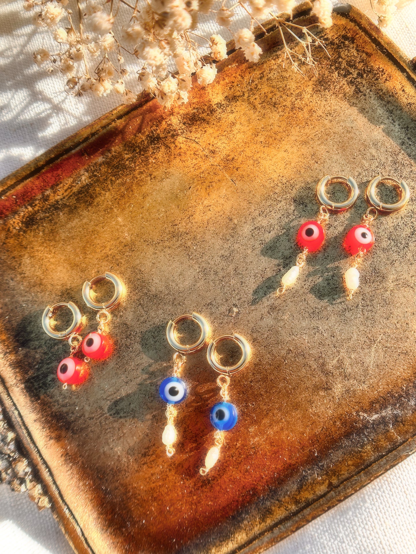 Pairs of Gold Stainless Steel earrings with Red and Blue Evil Eye beads with Faux Pearls on a stand and with a light blue background.