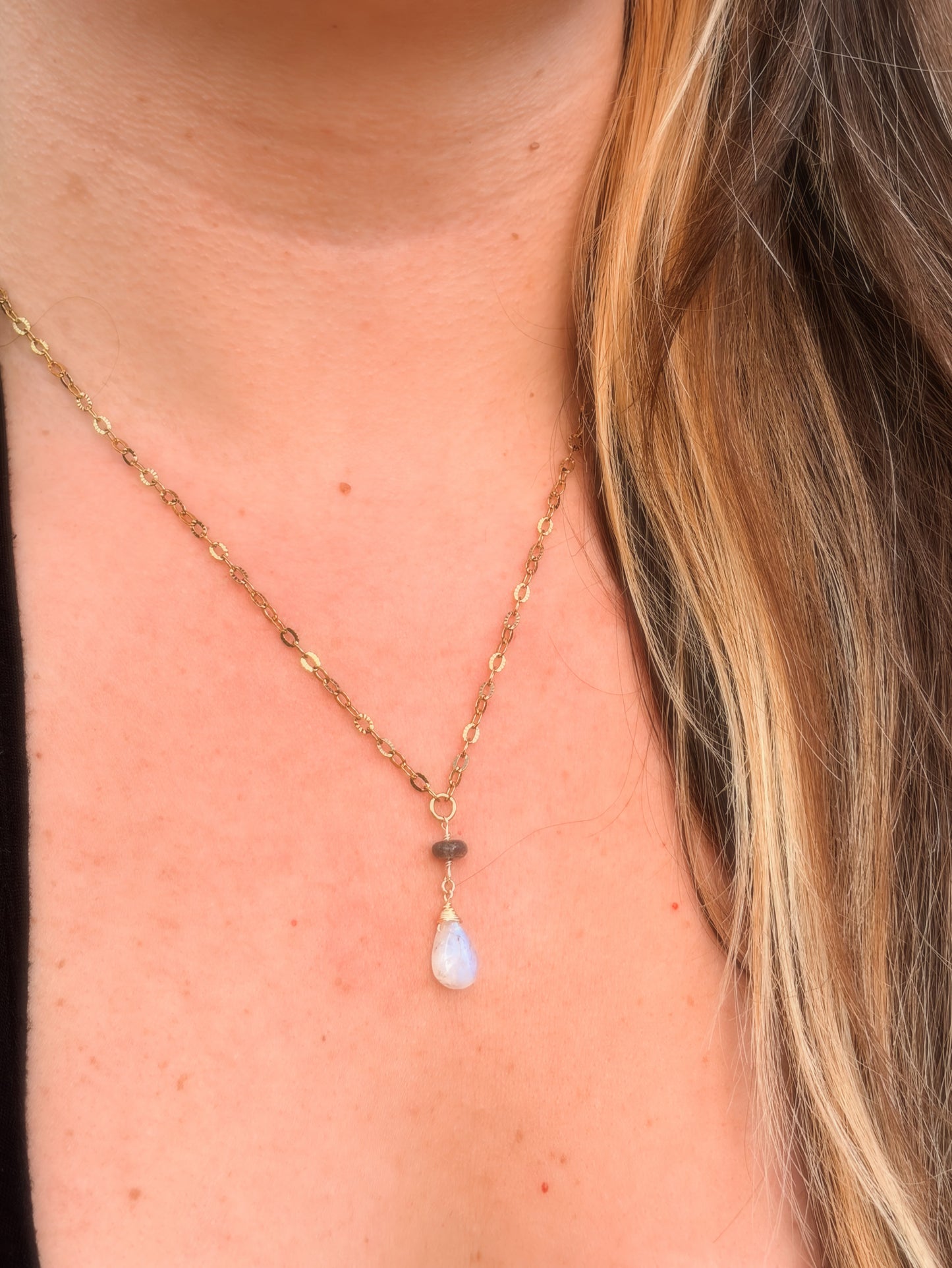 Close-up of a rainbow moonstone and labradorite stainless steel gold necklace with a teardrop pendant on a person wearing a black top.