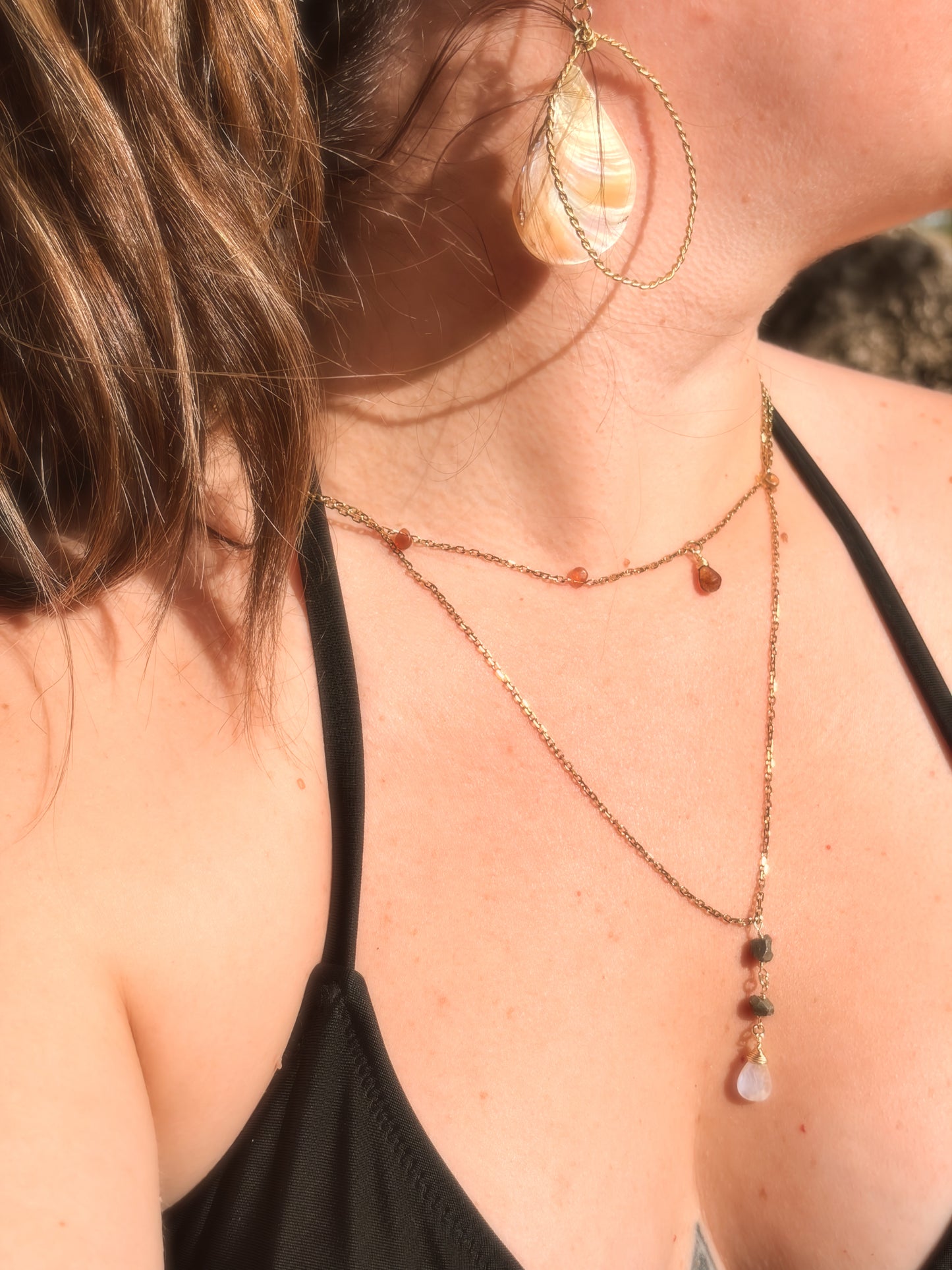 Handcrafted twisted gold brass hoop earrings with hanging pink and cream faux abalone shell charms displayed on a beach rock background.