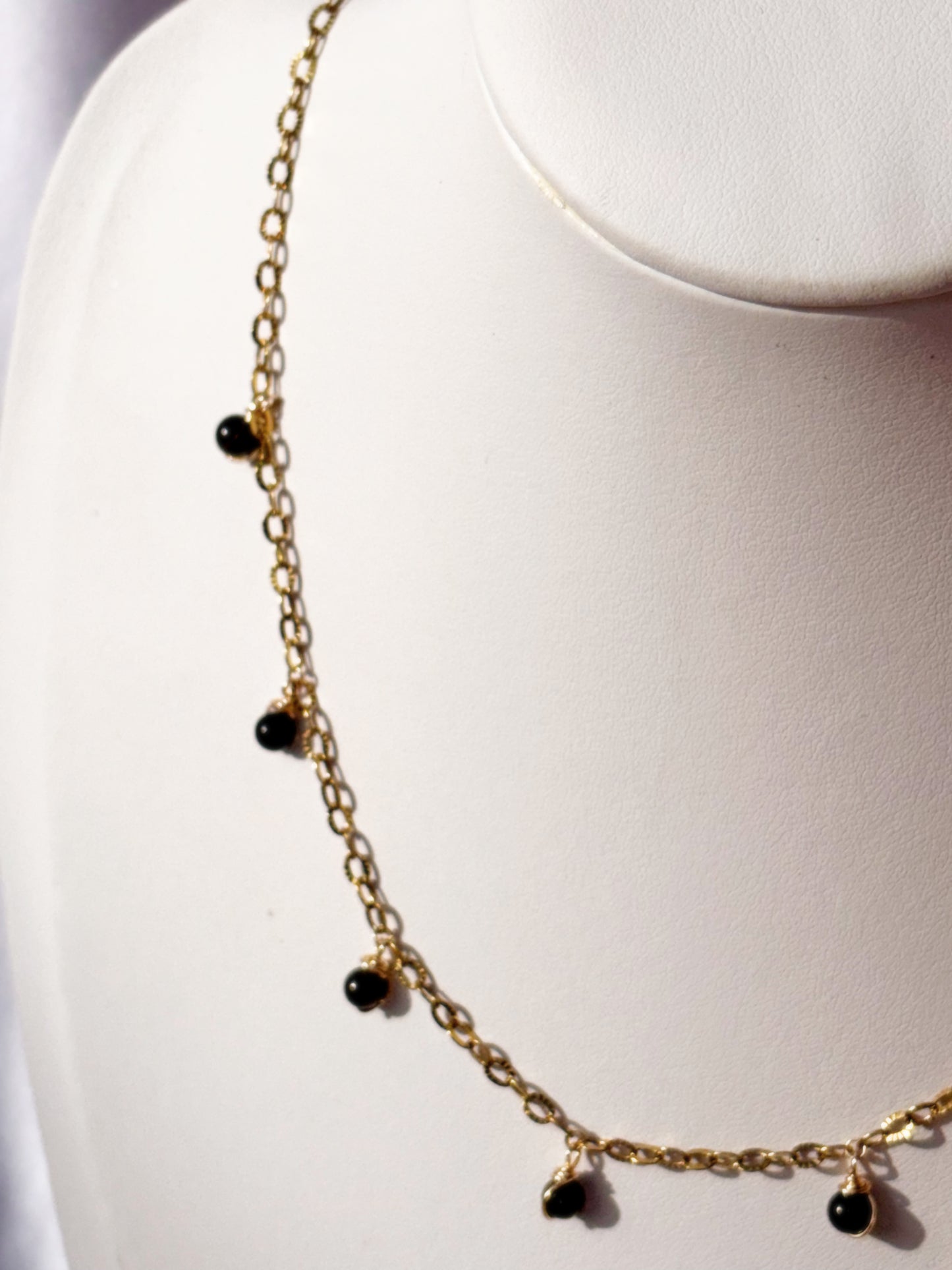 Close up of Gold Stainless Steel necklace with black gold sheen obsidian beads on a white stand against a light background.