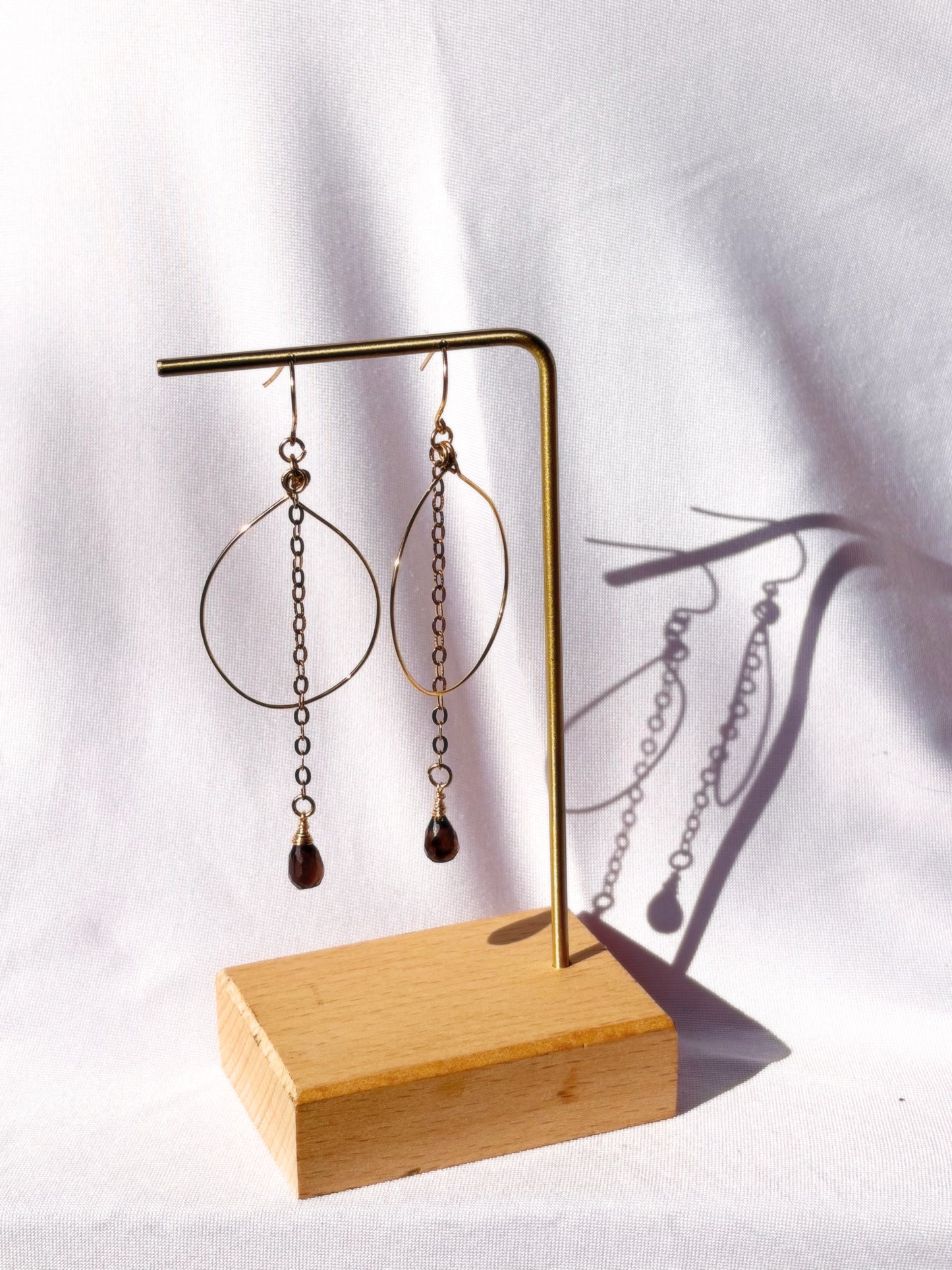A pair of gold-toned dangle earrings with large open hoop shapes, short chain links, and teardrop-shaped brown Smoky Qyartz bead drops, displayed on a gold stand against a light fabric background.