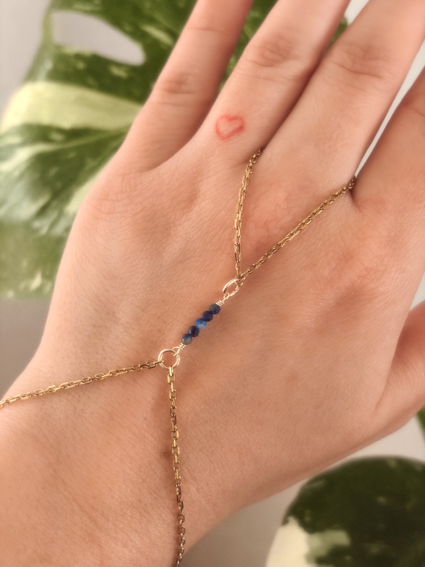 Gold Stainless Steel tarnish resistant handchain with blue Sodalite beads on a person's hand against a blurred background with a monsterra plant.
