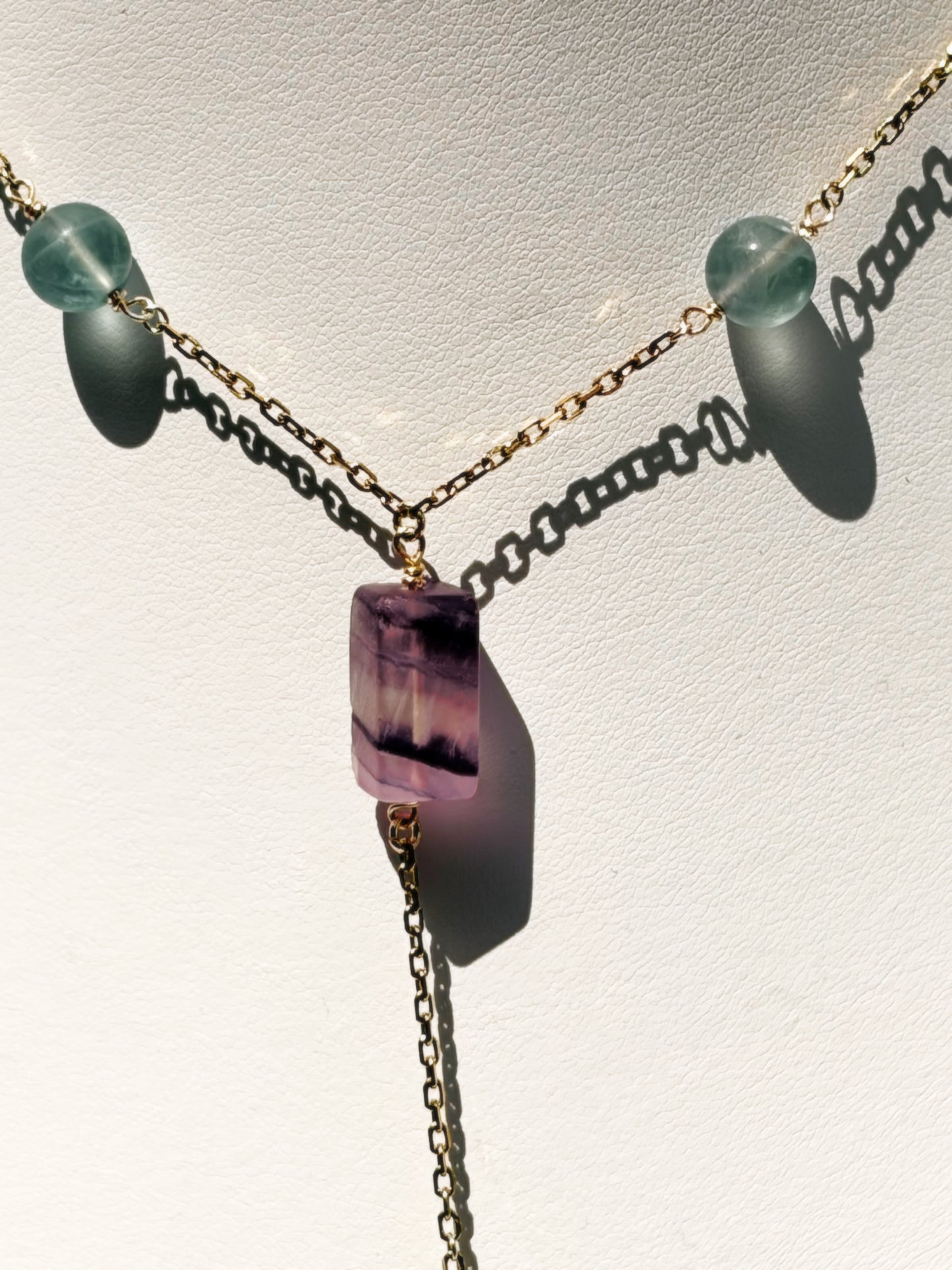 A delicate gold-tone lariat-style necklace displayed on a white jewelry bust. The necklace features a central faceted purple fluorite crystal pendant with small light fluorite blue-green beads on a stainless steel chain.