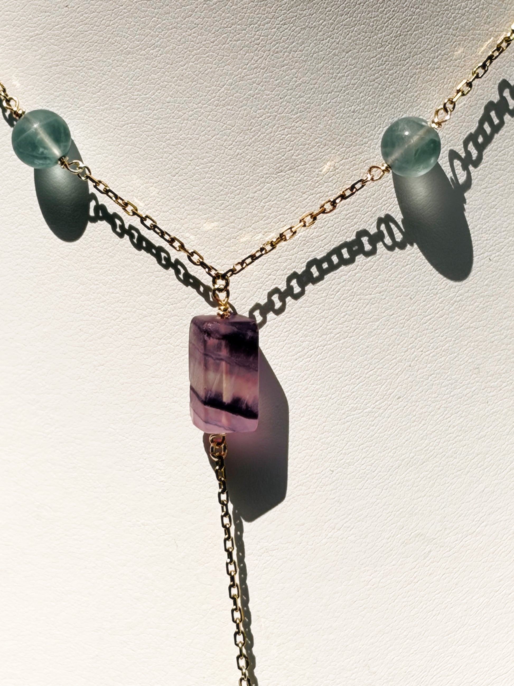 A delicate gold-tone lariat-style necklace displayed on a white jewelry bust. The necklace features a central faceted purple fluorite crystal pendant with small light fluorite blue-green beads on a stainless steel chain.