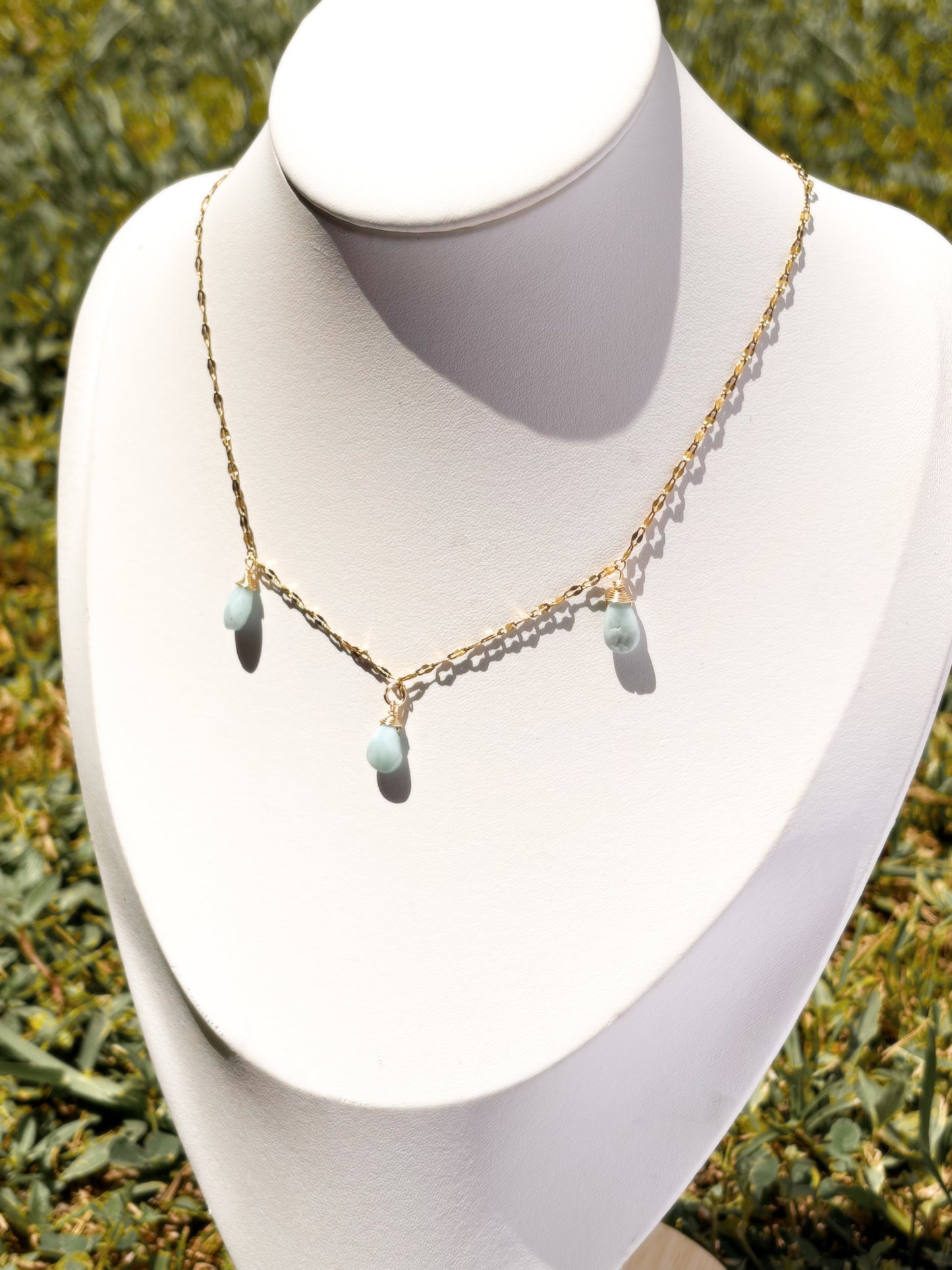 A delicate gold chain necklace with three small light blue teardrop Larimar beads, displayed on a white jewelry bust.