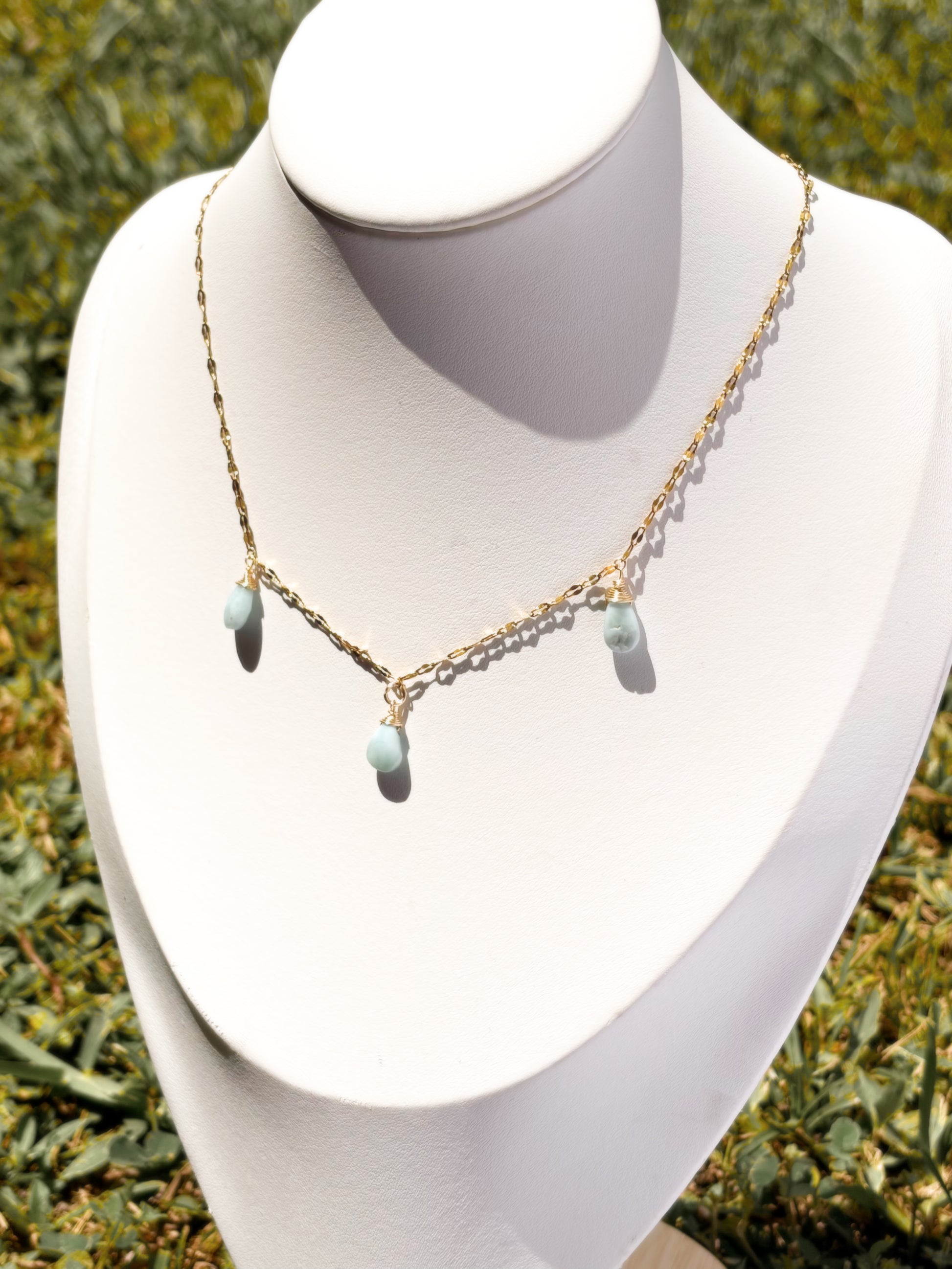 A delicate gold chain necklace with three small light blue teardrop Larimar beads, displayed on a white jewelry bust.
