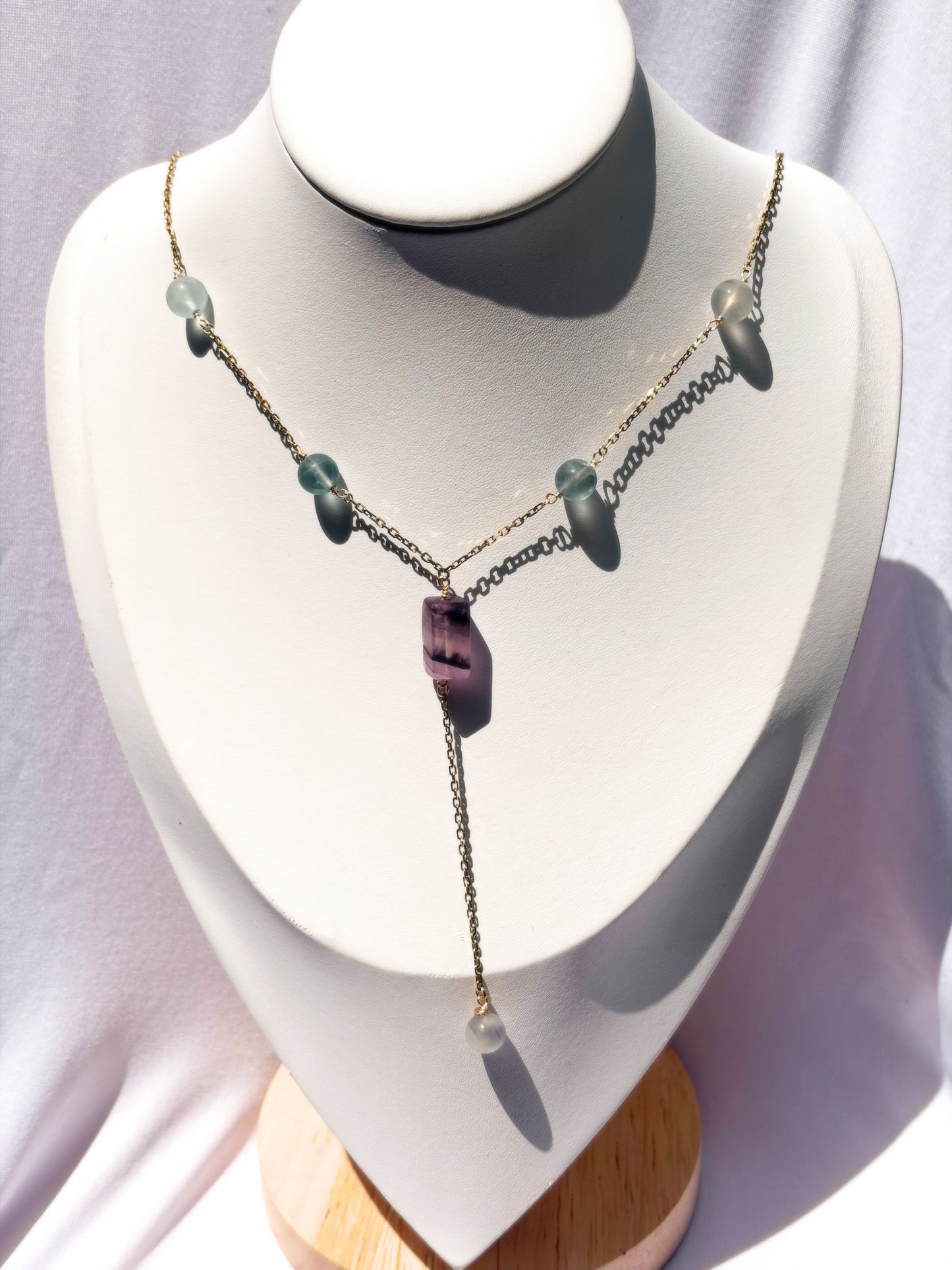 A delicate gold-tone lariat-style necklace displayed on a white jewelry bust. The necklace features a central faceted purple fluorite crystal pendant with small light fluorite blue-green beads on a stainless steel chain.