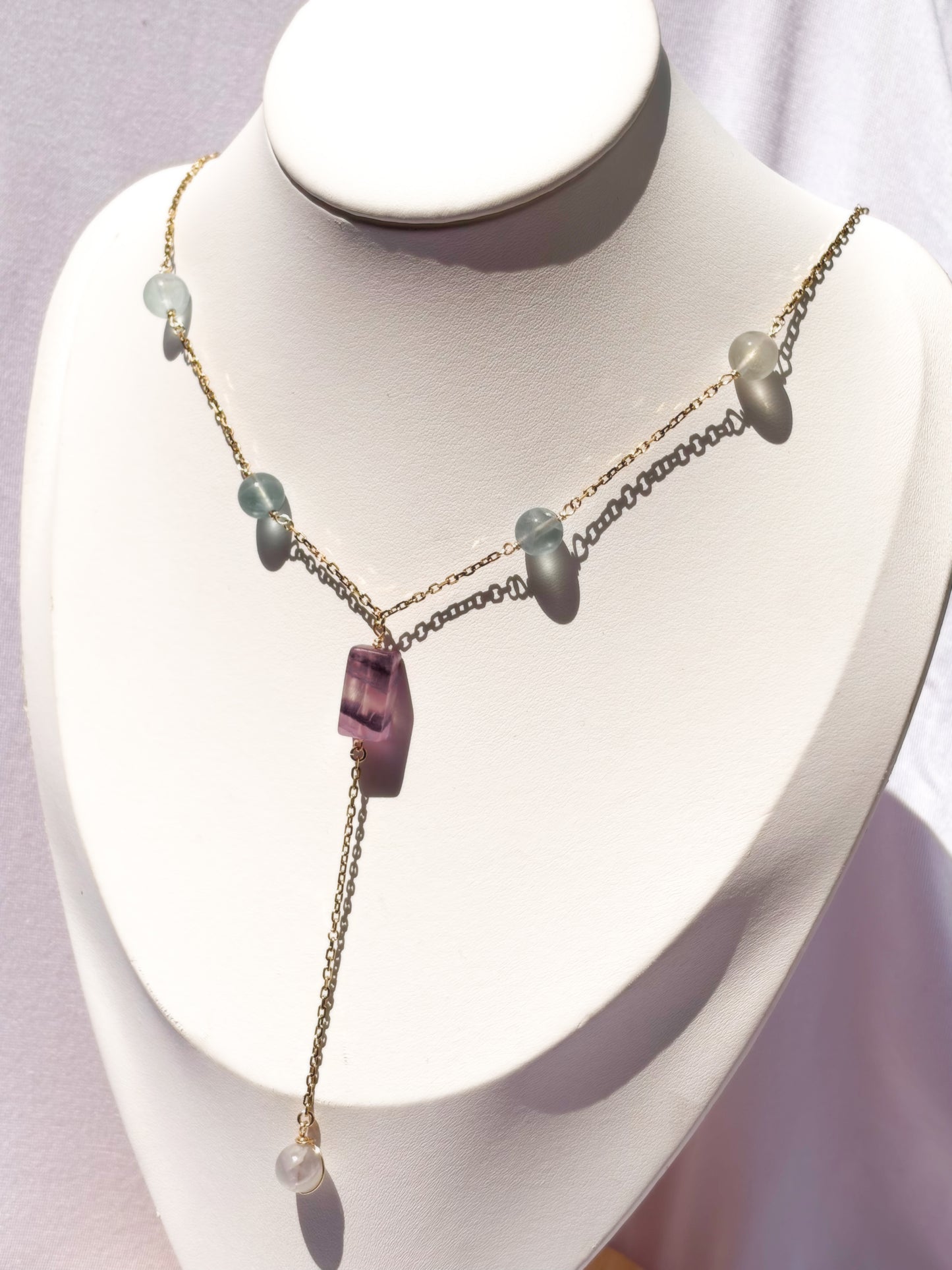 A delicate gold-tone lariat-style necklace displayed on a white jewelry bust. The necklace features a central faceted purple fluorite crystal pendant with small light fluorite blue-green beads on a stainless steel chain.