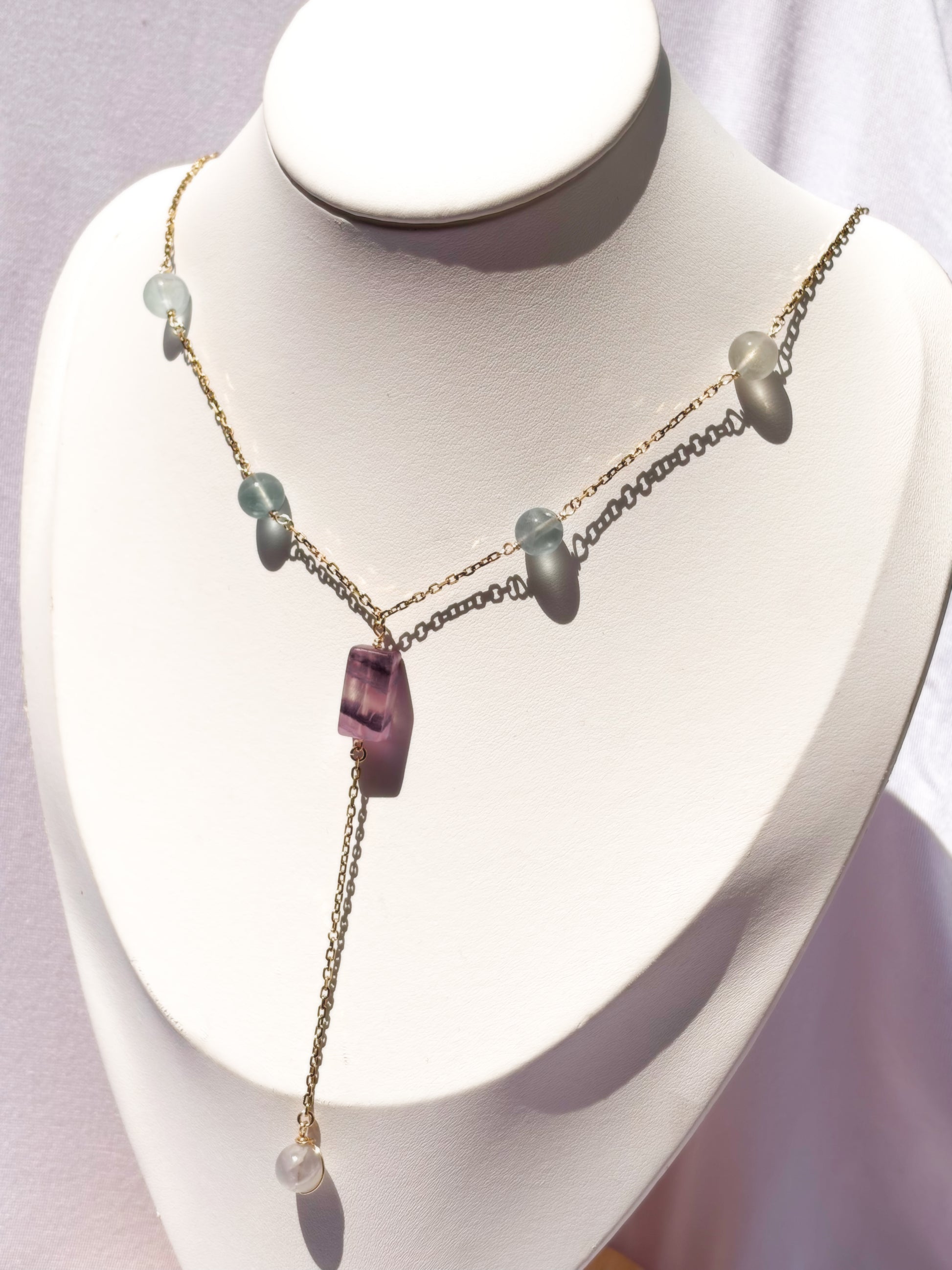 A delicate gold-tone lariat-style necklace displayed on a white jewelry bust. The necklace features a central faceted purple fluorite crystal pendant with small light fluorite blue-green beads on a stainless steel chain.