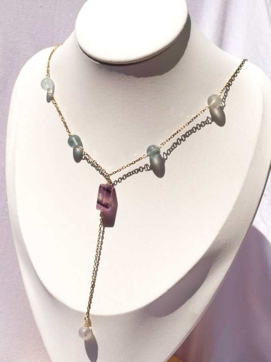 A delicate gold-tone lariat-style necklace displayed on a white jewelry bust. The necklace features a central faceted purple fluorite crystal pendant with small light fluorite blue-green beads on a stainless steel chain.