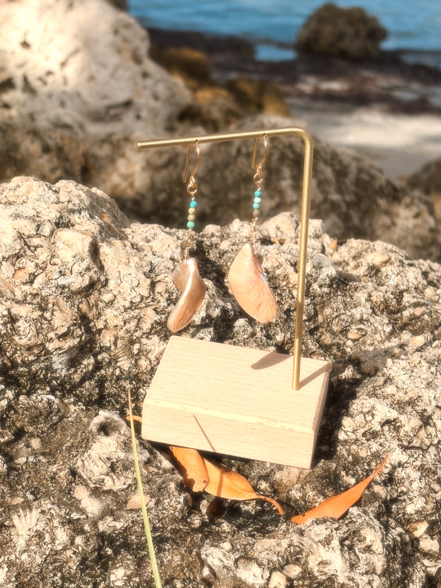 Handmade brass earrings with dangling pearlescent faux abalone shell charms and turquoise on a coastal rock backdrop.