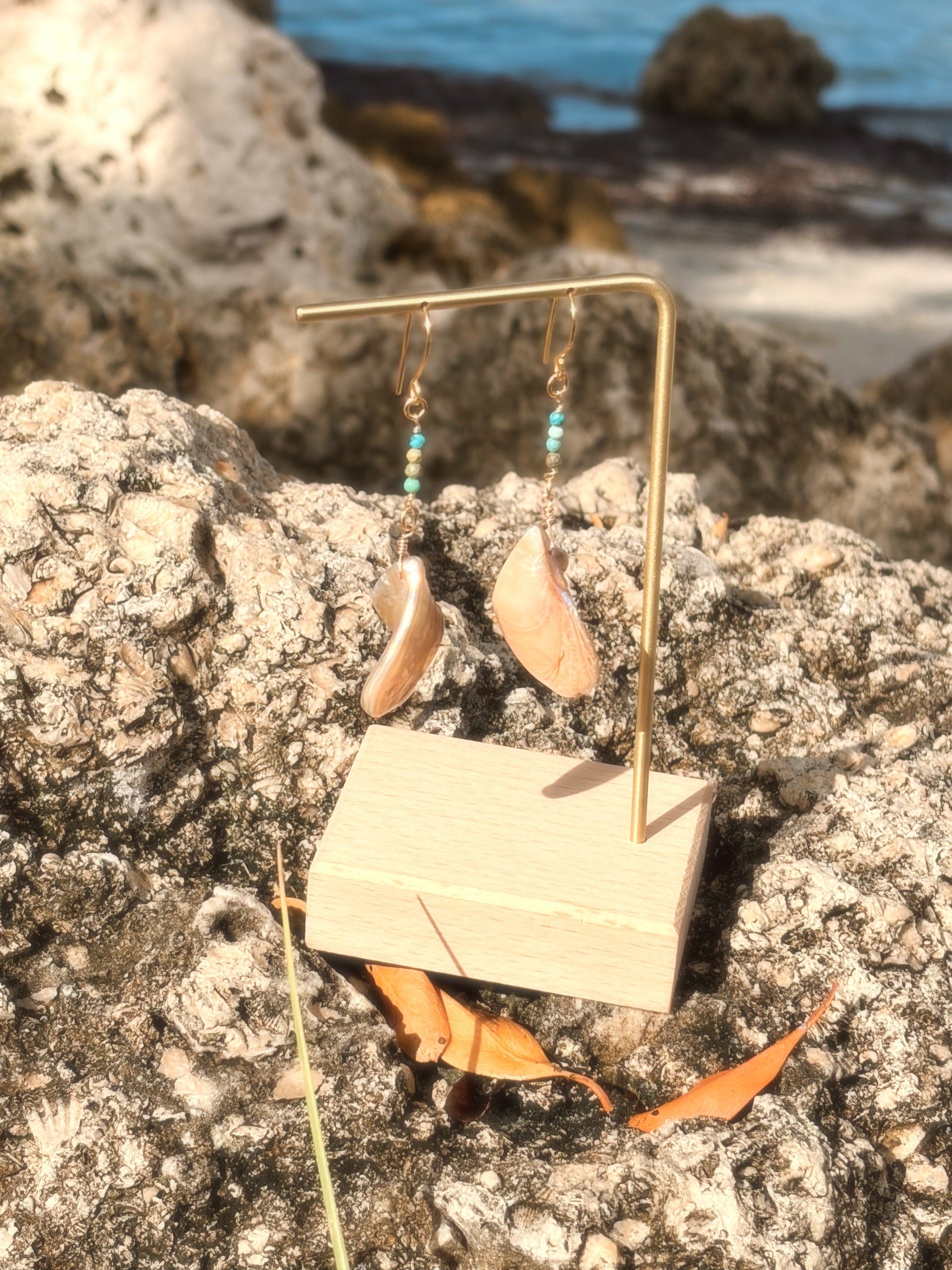Handmade brass earrings with dangling pearlescent faux abalone shell charms and turquoise on a coastal rock backdrop.