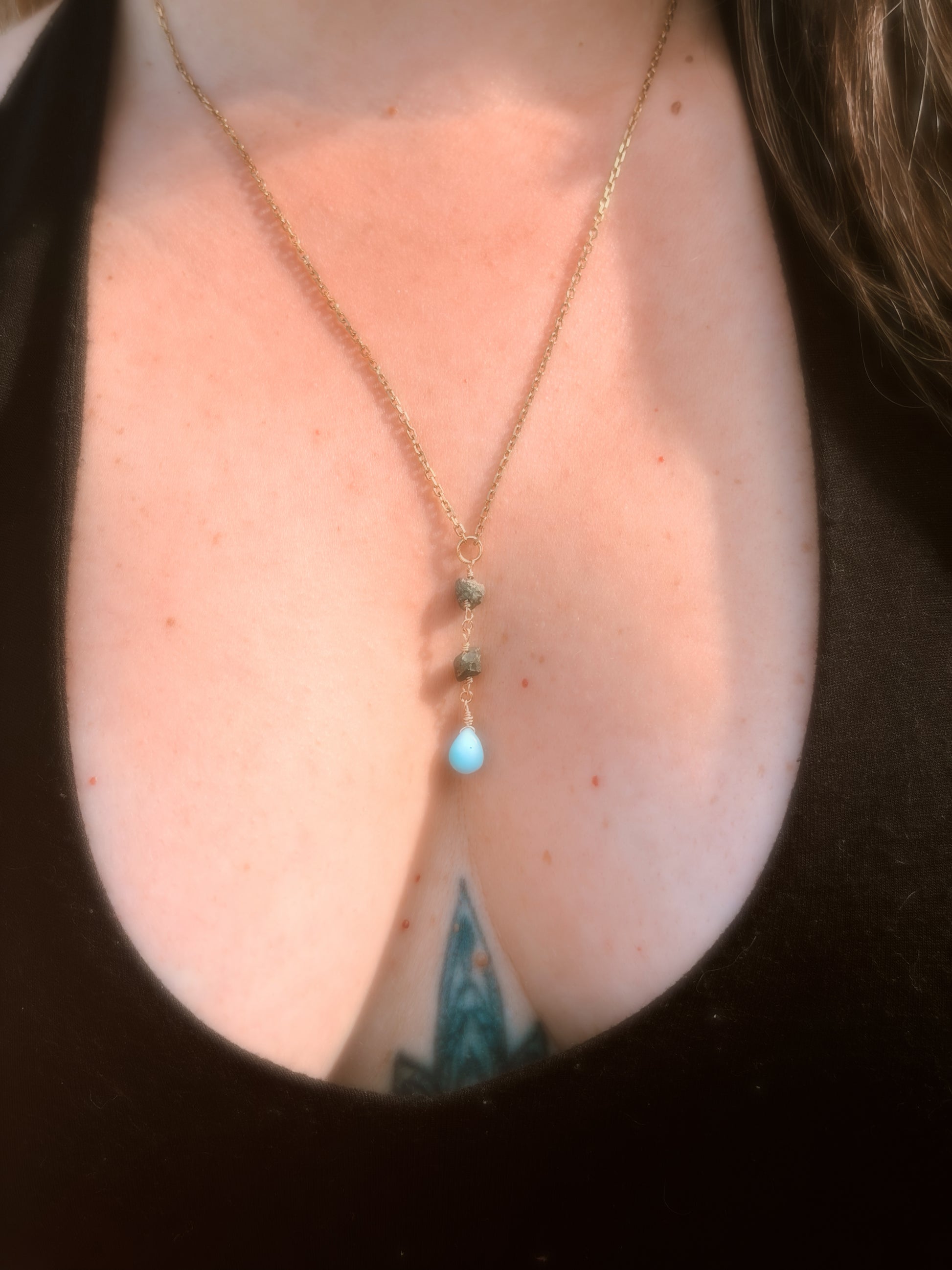 Close-up of a person wearing a delicate gold stainless steel necklace with Pyrite and Larimar beads on a plain background wearing a black top.