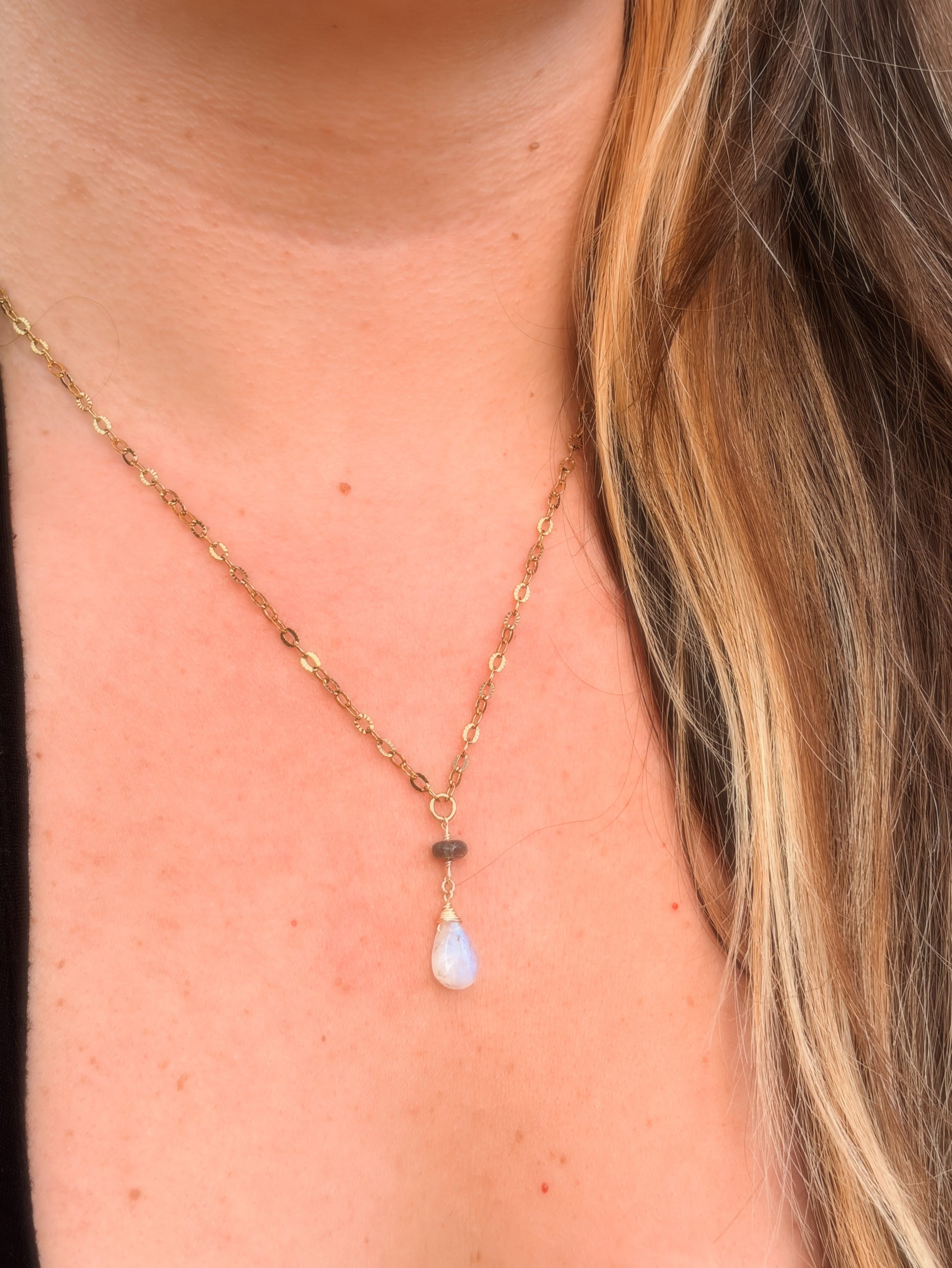 Close-up of a rainbow moonstone and labradorite stainless steel gold necklace with a teardrop pendant on a person wearing a black top.