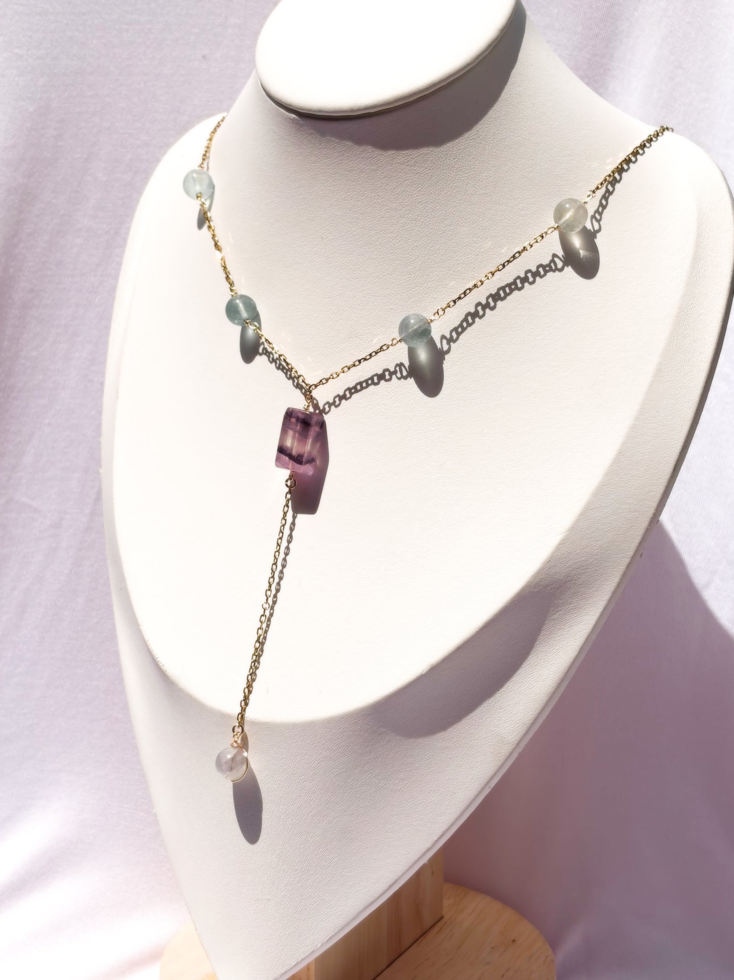 A delicate gold-tone lariat-style necklace displayed on a white jewelry bust. The necklace features a central faceted purple fluorite crystal pendant with small light fluorite blue-green beads on a stainless steel chain.