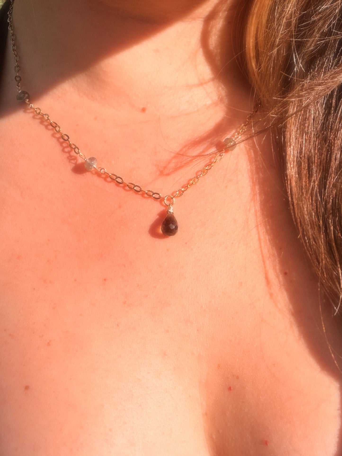 Close-up of a smoky quartz and labradorite stainless steel gold necklace with a teardrop pendant on a person wearing a black top.