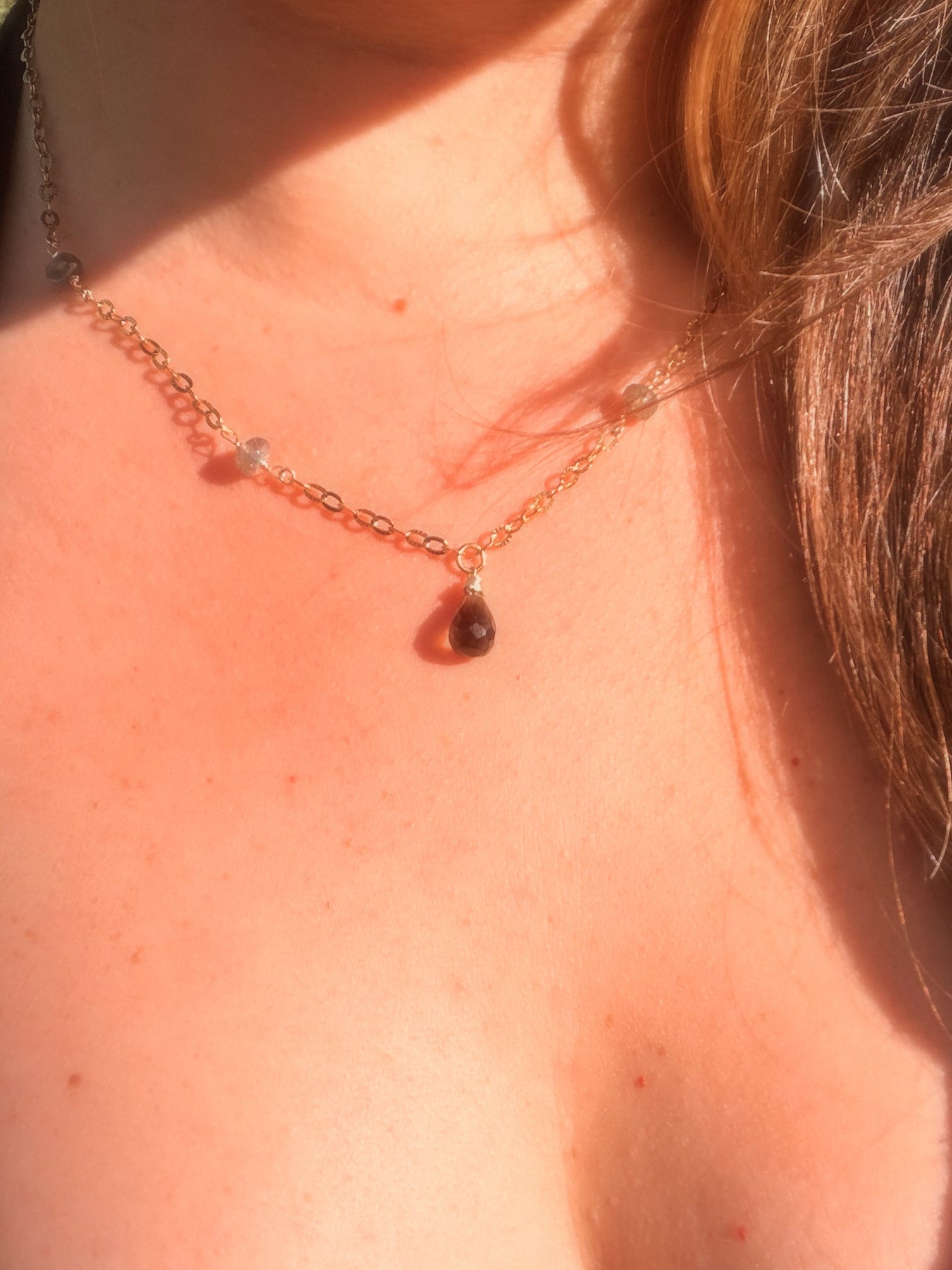 Close-up of a smoky quartz and labradorite stainless steel gold necklace with a teardrop pendant on a person wearing a black top.