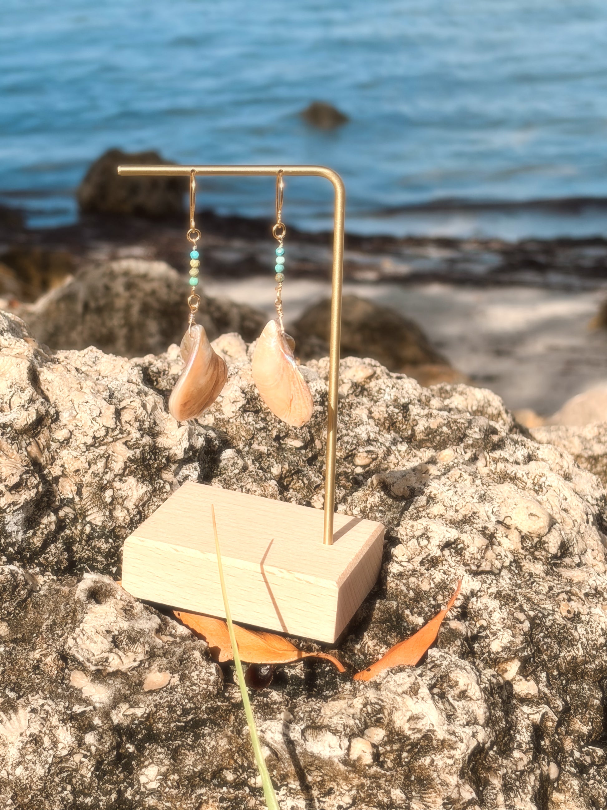 Handmade brass earrings with dangling pearlescent faux abalone shell charms and turquoise on a coastal rock backdrop.