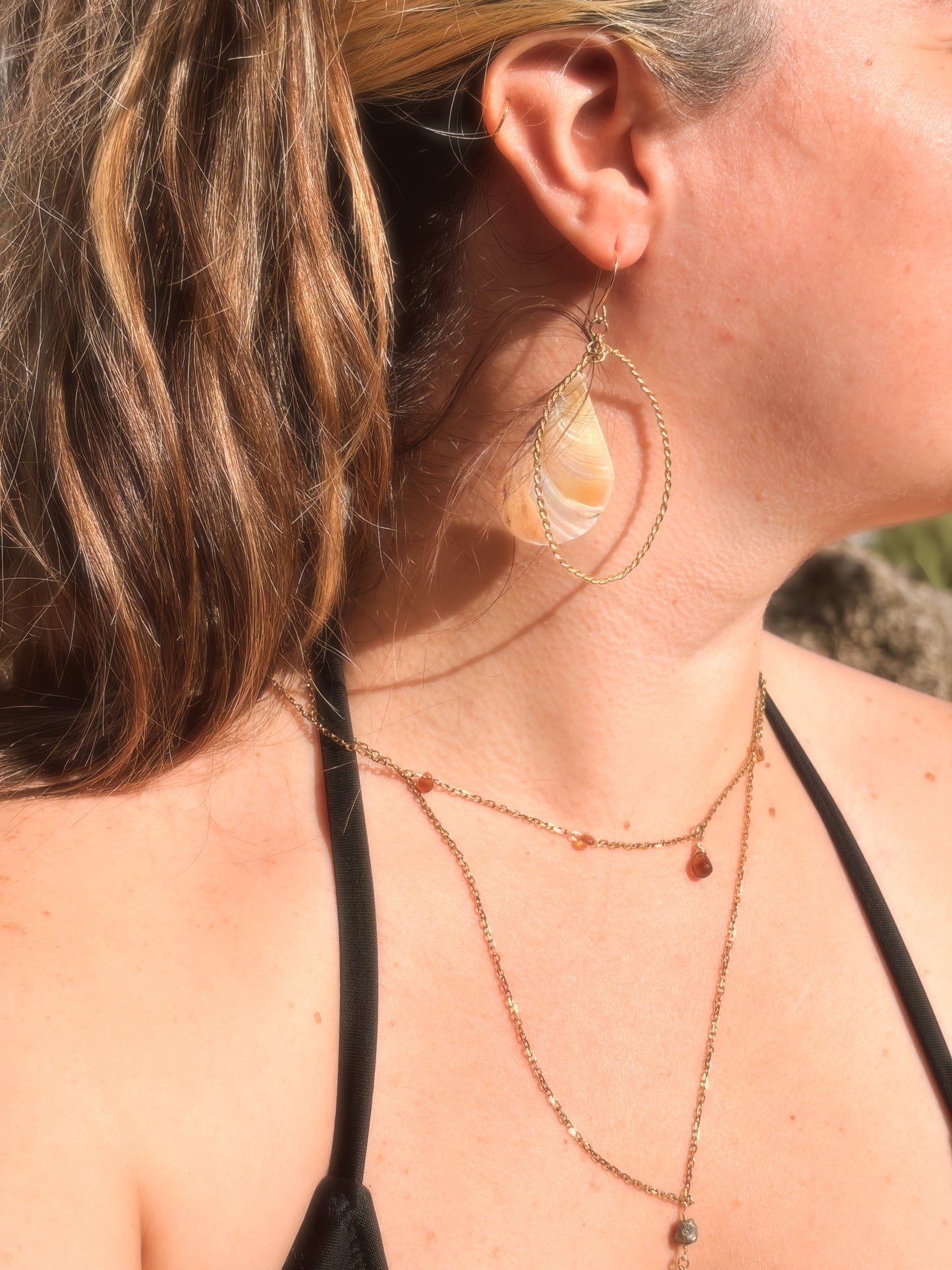 Handcrafted twisted gold brass hoop earrings with hanging pink and cream faux abalone shell charms displayed on a beach rock background.