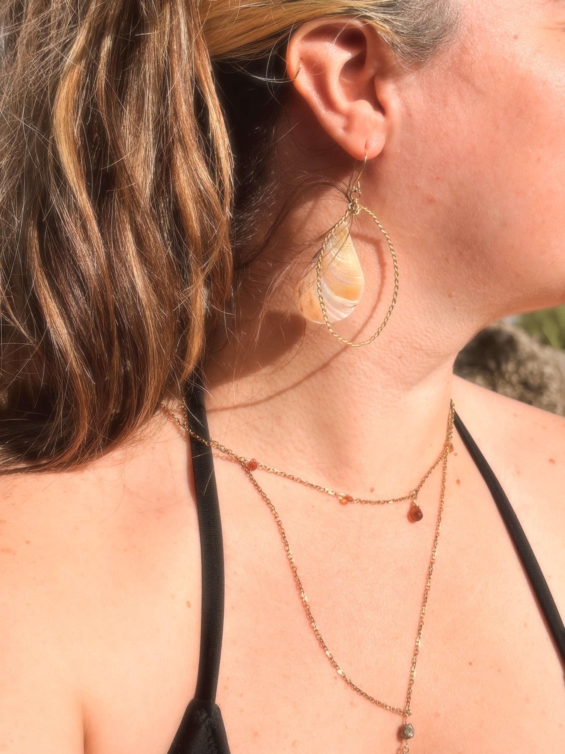 Handcrafted twisted gold brass hoop earrings with hanging pink and cream faux abalone shell charms displayed on a beach rock background.