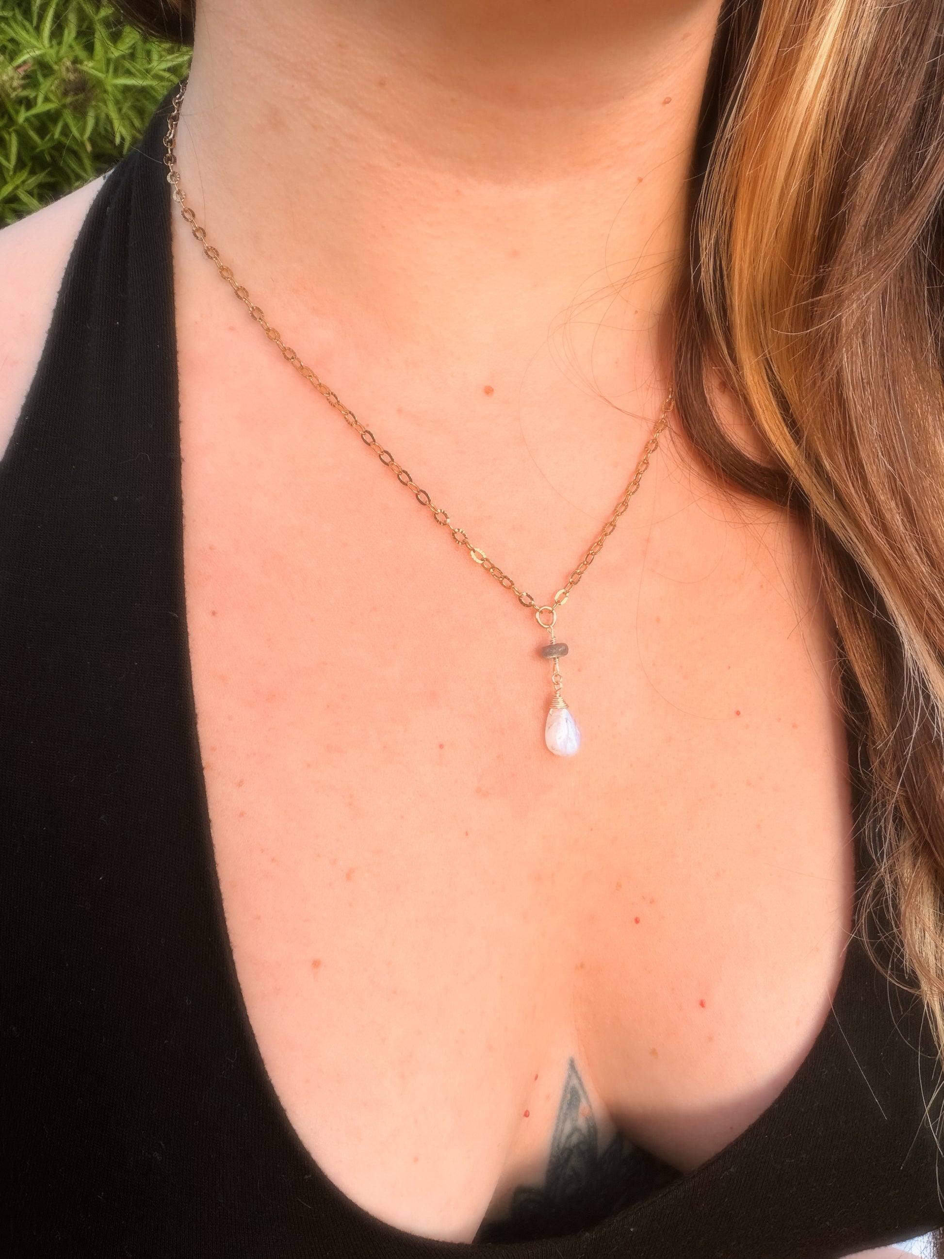 Close-up of a rainbow moonstone and labradorite stainless steel gold necklace with a teardrop pendant on a person wearing a black top.