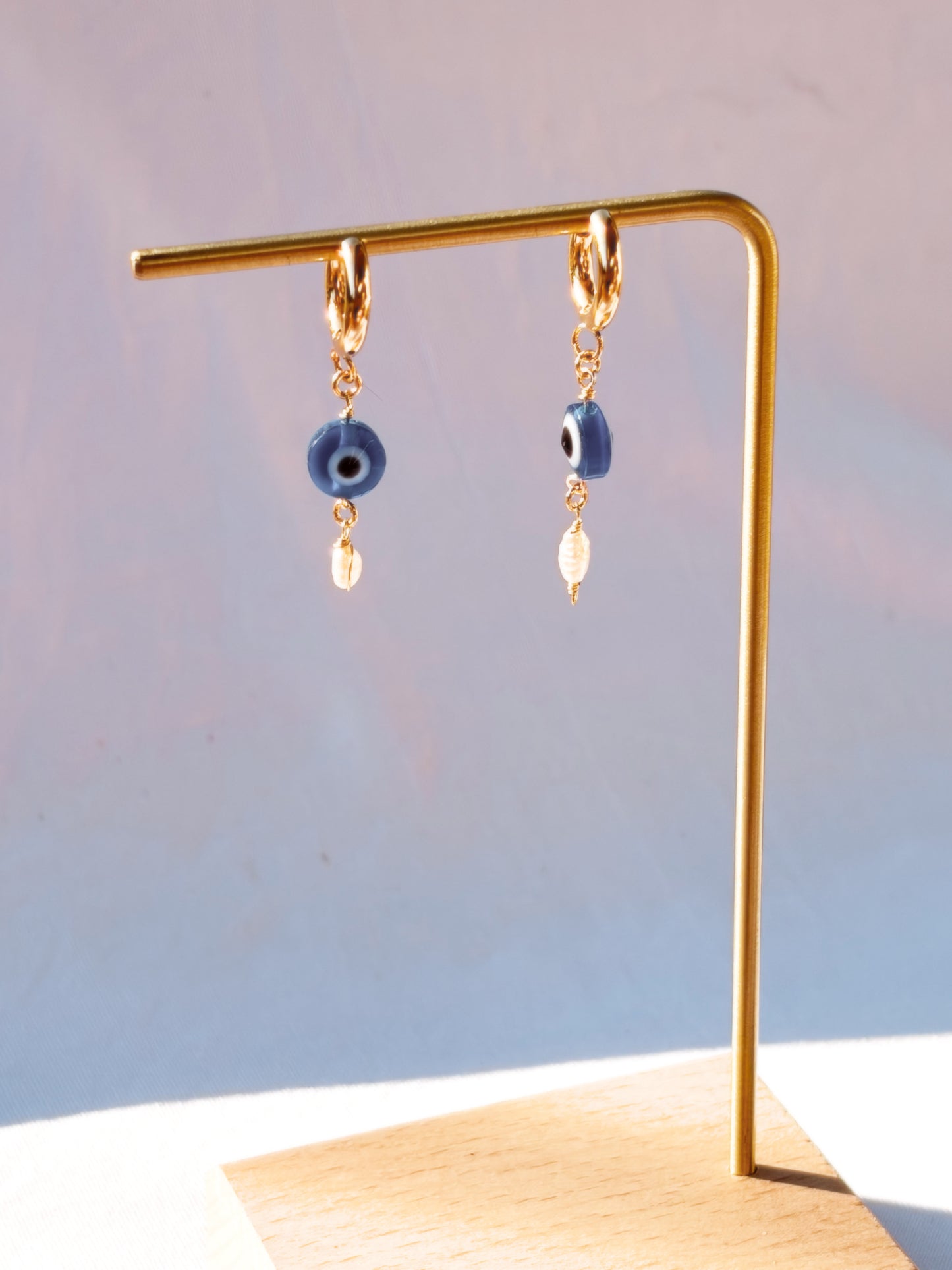 Gold Stainless Steel earrings with Blue Evil Eye beads with Faux Pearls on a stand and with a light blue background.