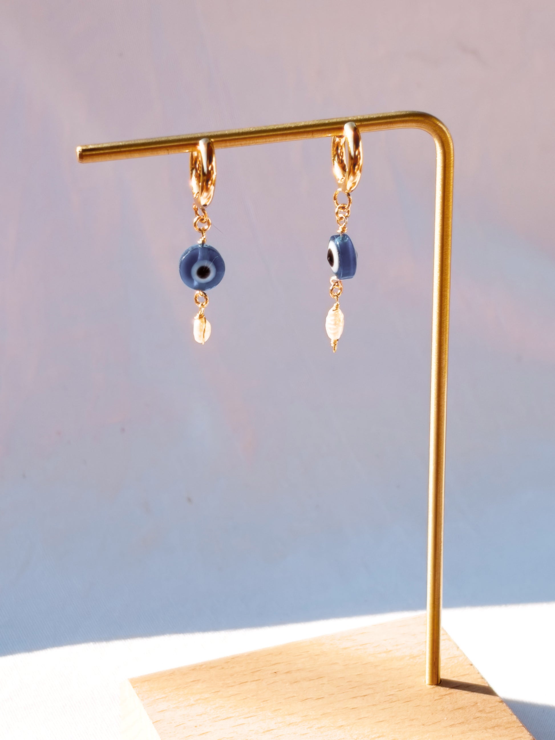 Gold Stainless Steel earrings with Blue Evil Eye beads with Faux Pearls on a stand and with a light blue background.