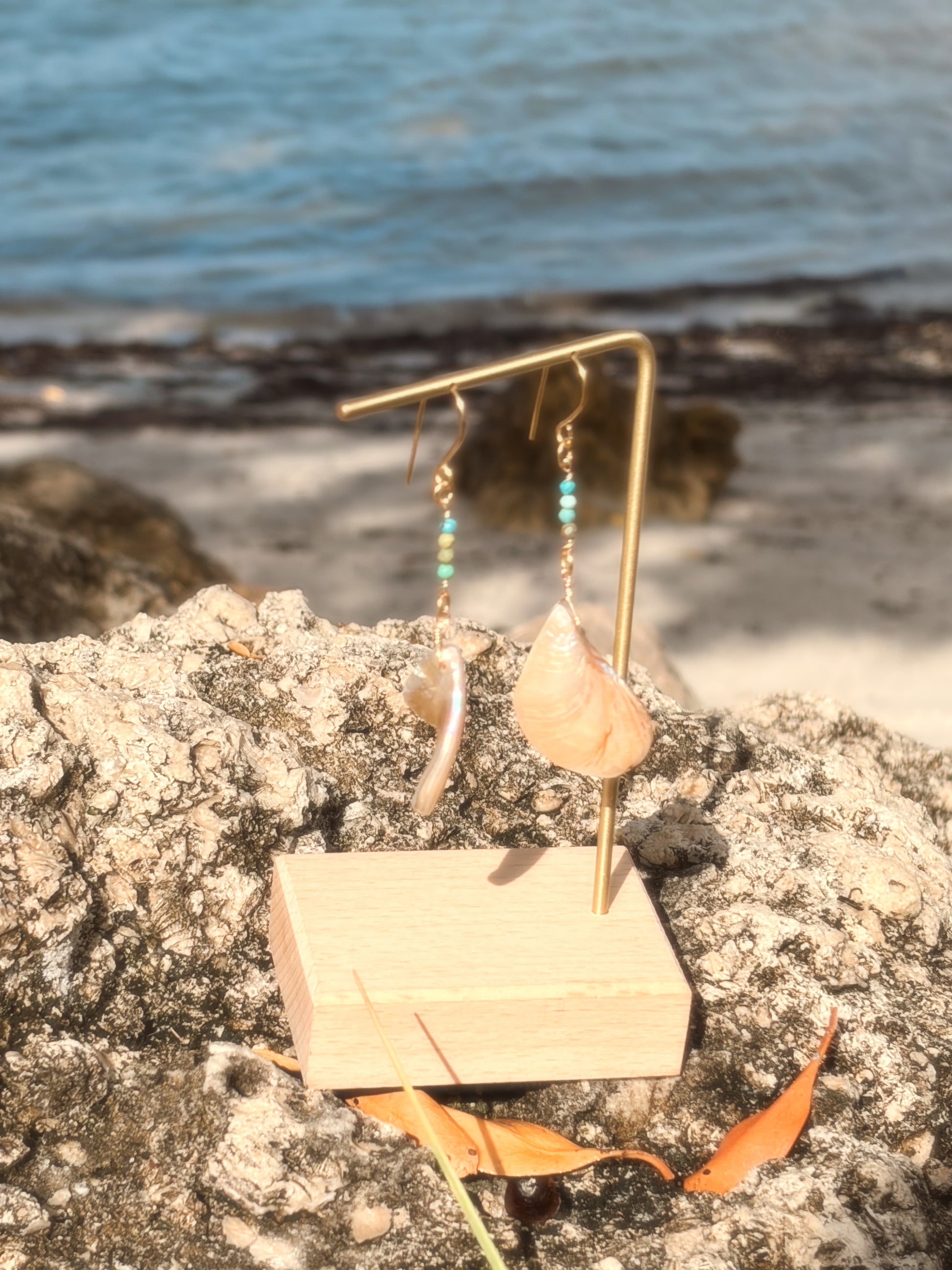 Handmade brass earrings with dangling pearlescent faux abalone shell charms and turquoise on a coastal rock backdrop.
