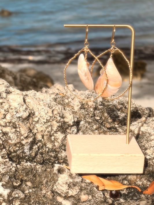 Pair of wire wrapped hoop shell gold earrings on a stand with a beach background