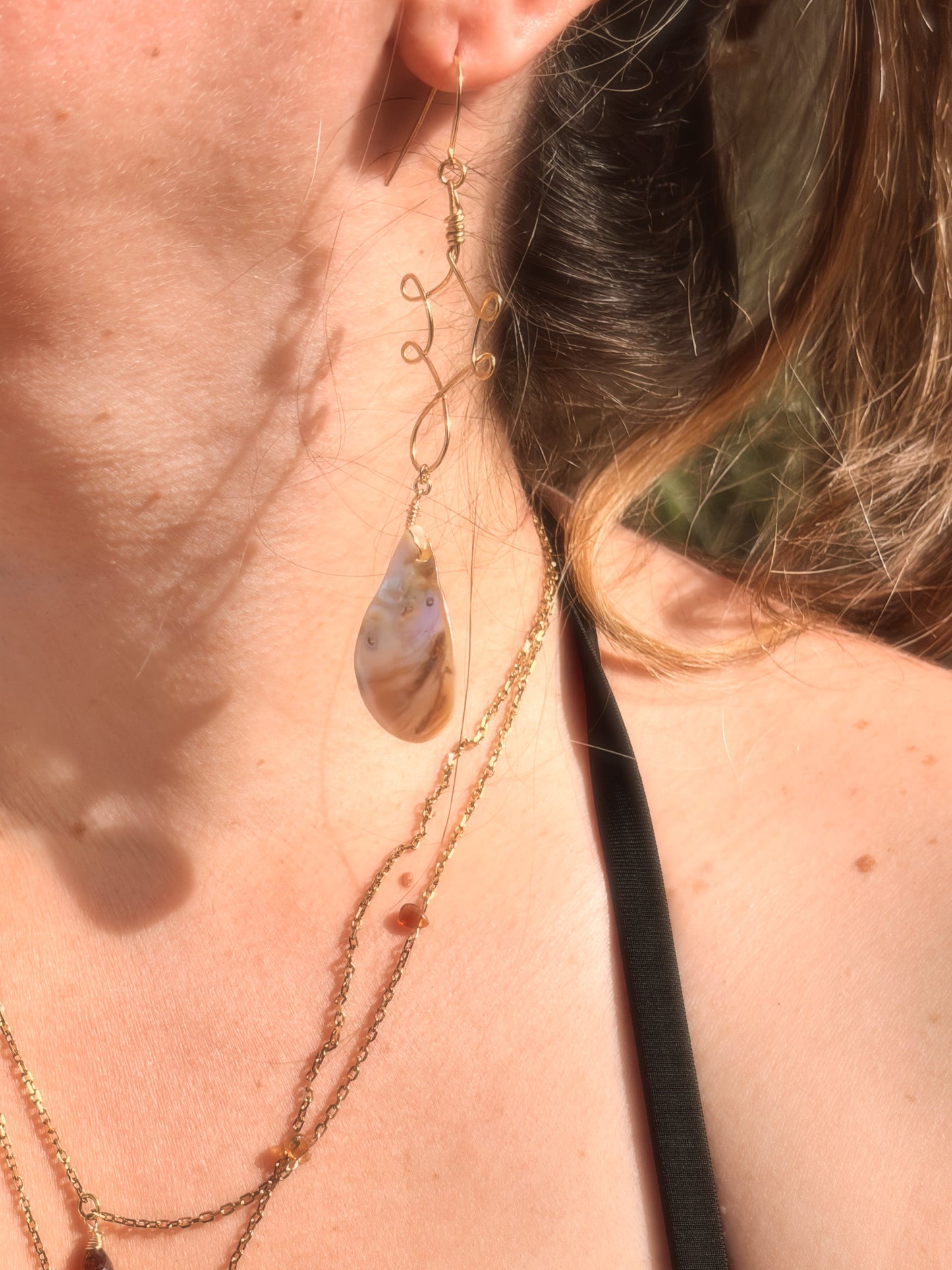 Close-up of a person wearing wire wrapped stainless steel gold brass earrings with an abalone shell bead and layered necklaces with a blurred background