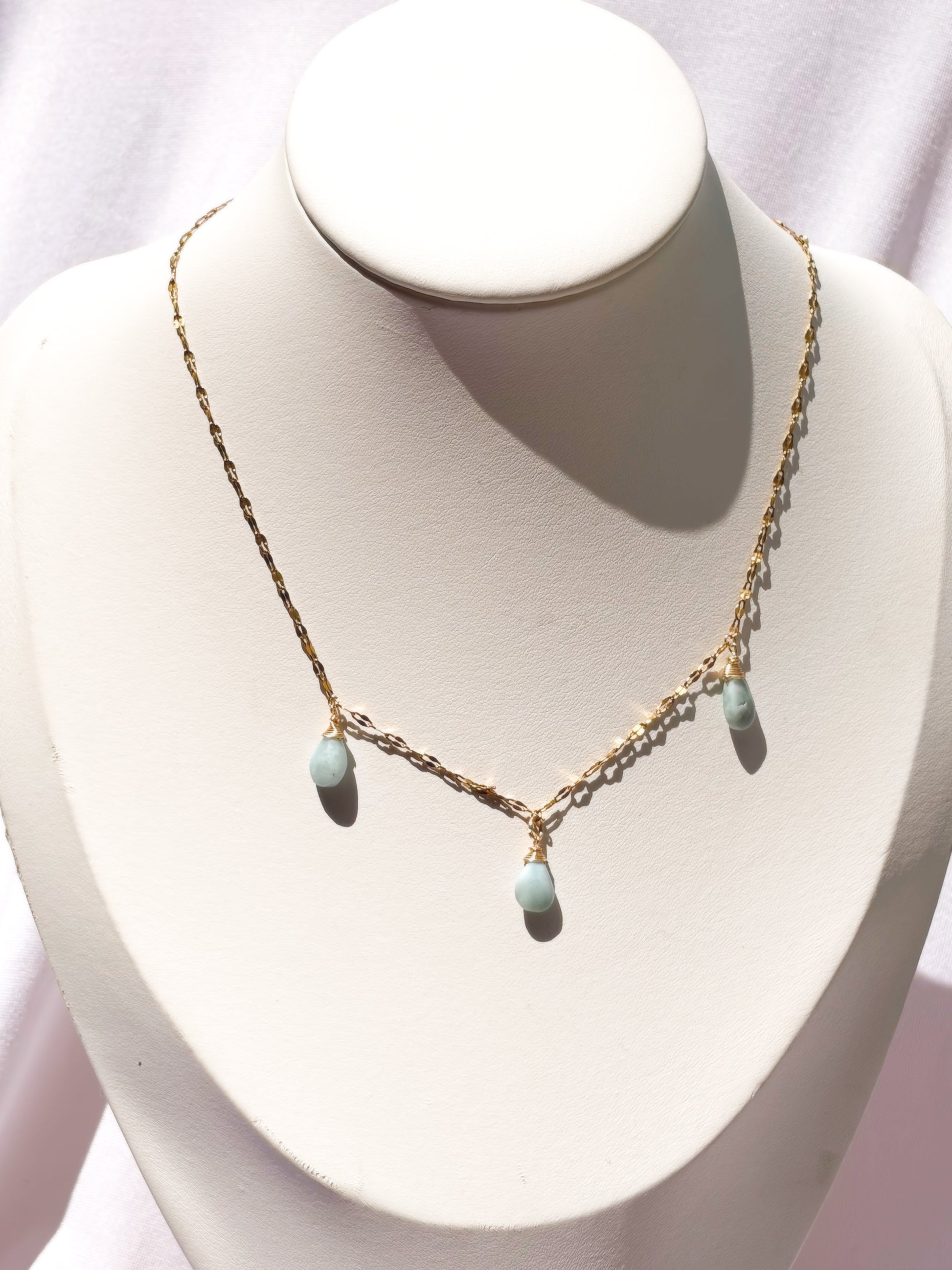 A delicate gold chain necklace with three small light blue teardrop Larimar beads, displayed on a white jewelry bust.