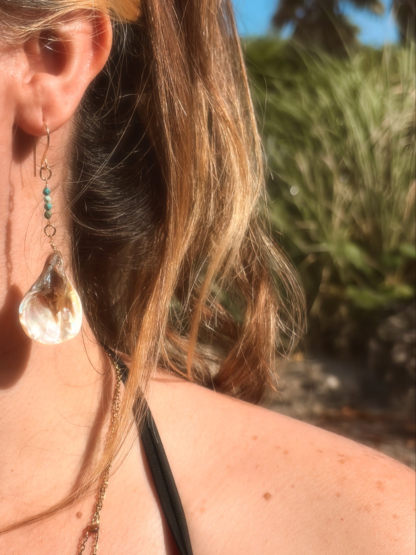Handmade brass earrings with dangling pearlescent faux abalone shell charms and turquoise on a coastal rock backdrop.