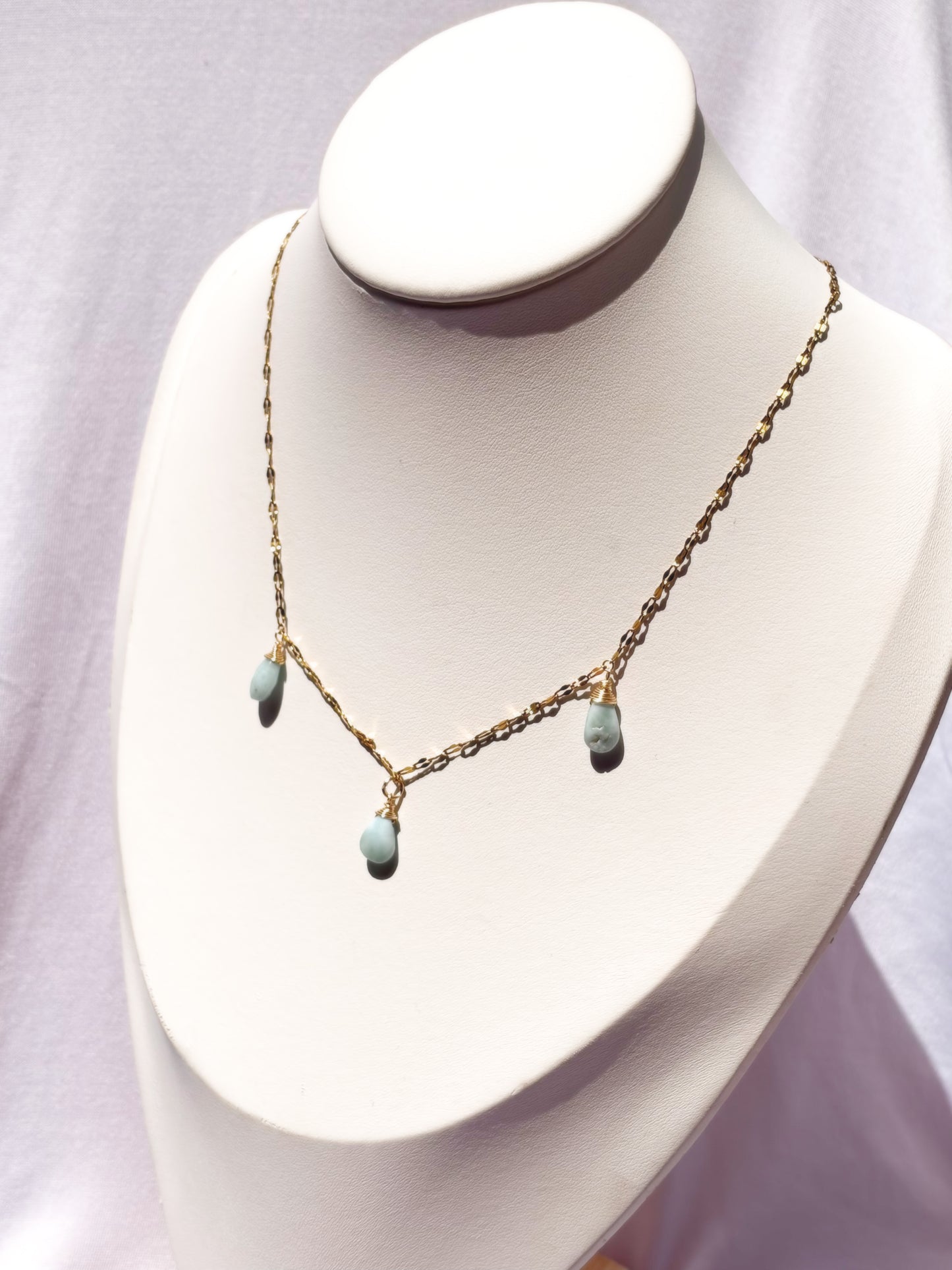 A delicate gold chain necklace with three small light blue teardrop Larimar beads, displayed on a white jewelry bust.