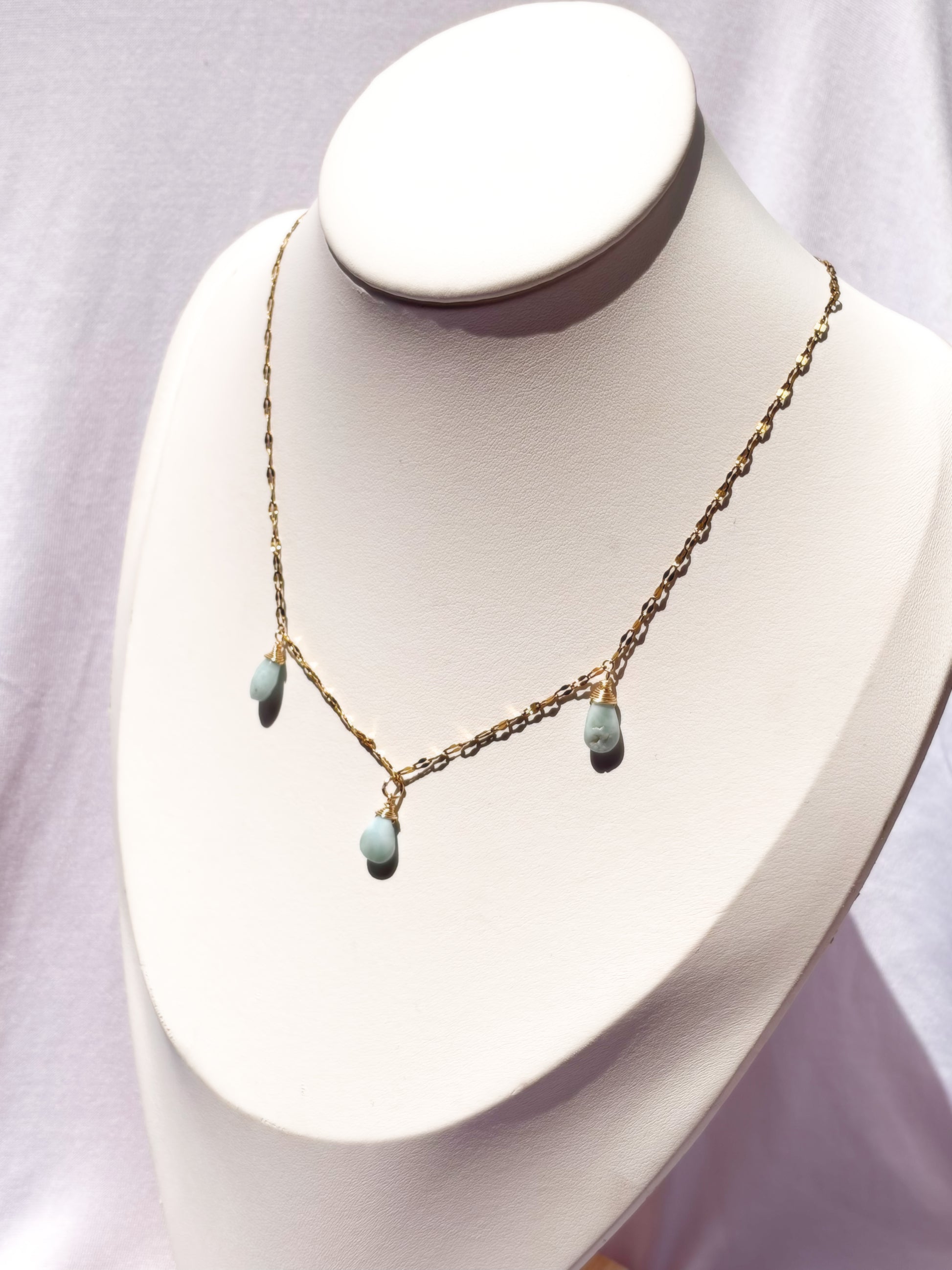 A delicate gold chain necklace with three small light blue teardrop Larimar beads, displayed on a white jewelry bust.