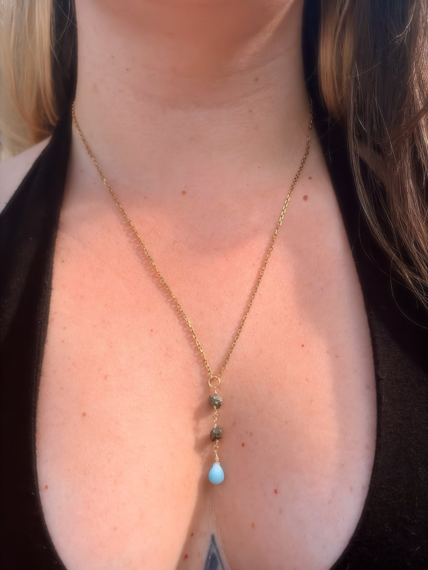 Close-up of a person wearing a delicate gold stainless steel necklace with Pyrite and Larimar beads on a plain background wearing a black top.