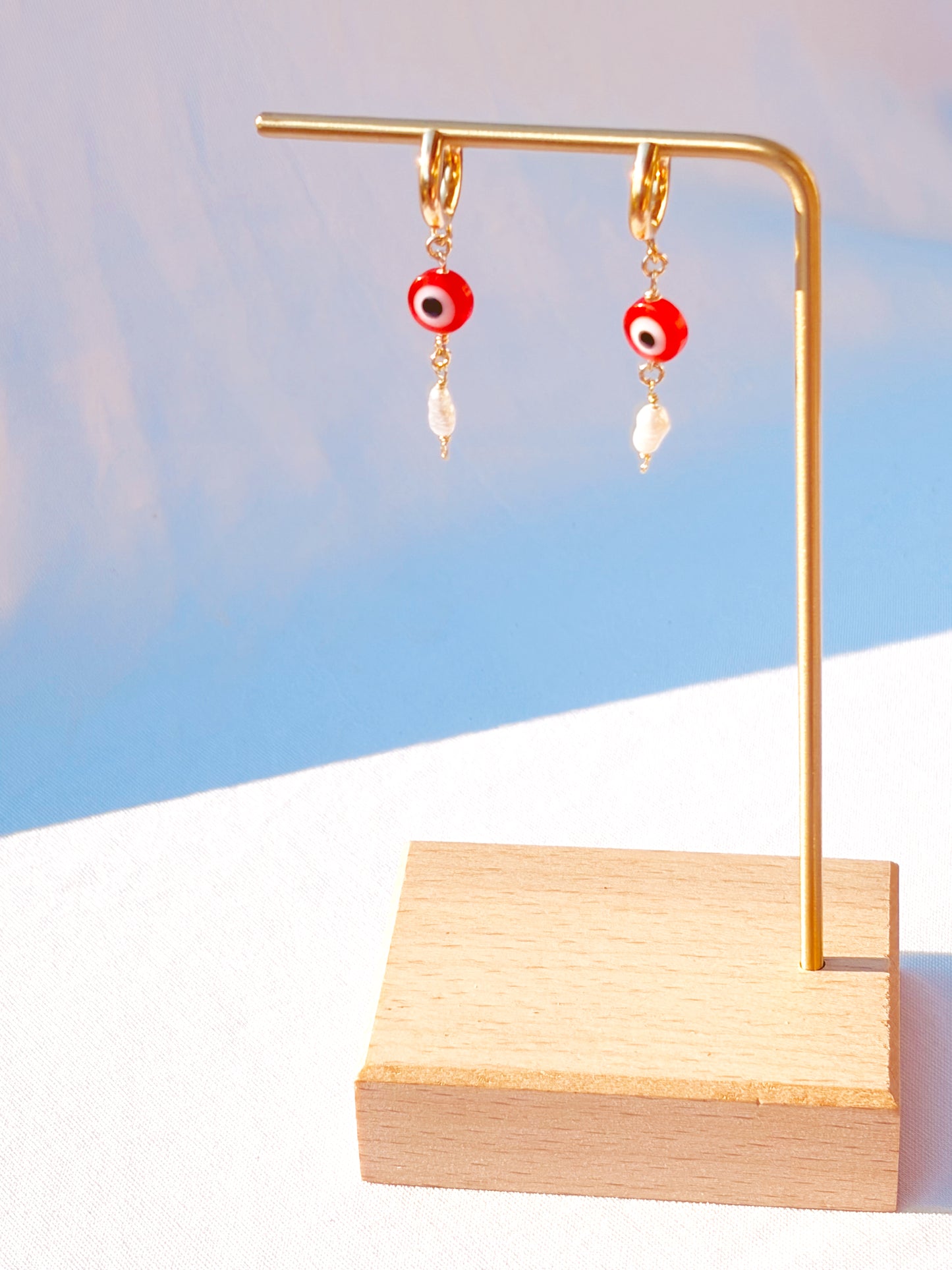 Gold Stainless Steel earrings with Red Evil Eye beads with Faux Pearls on a stand and with a light blue background.