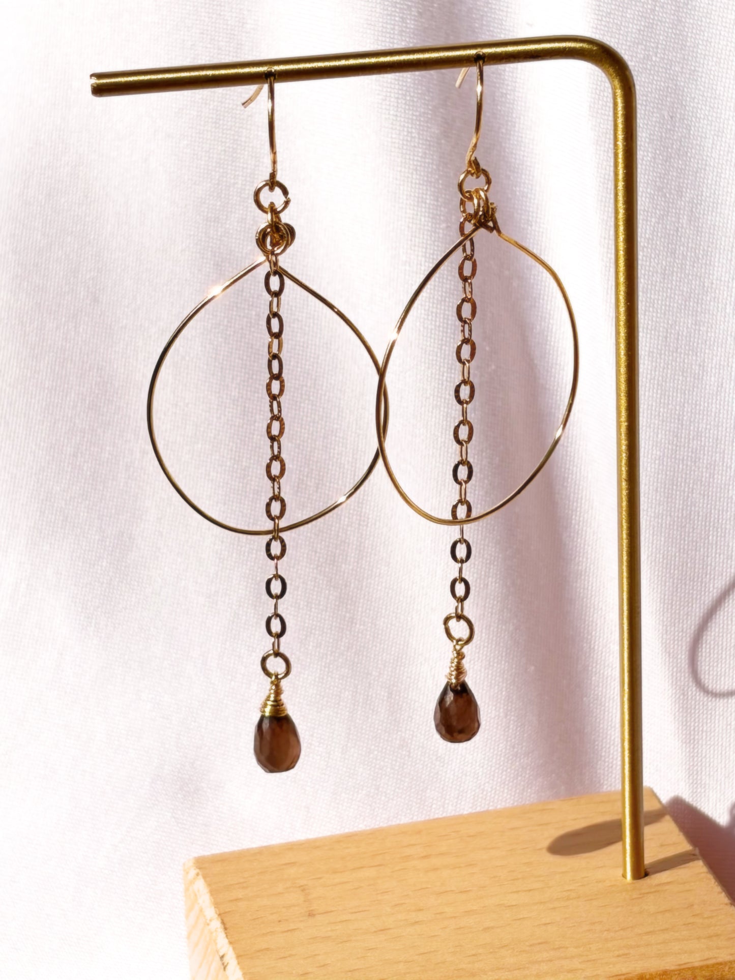 A pair of gold-toned dangle earrings with large open hoop shapes, short chain links, and teardrop-shaped brown Smoky Qyartz bead drops, displayed on a gold stand against a light fabric background.