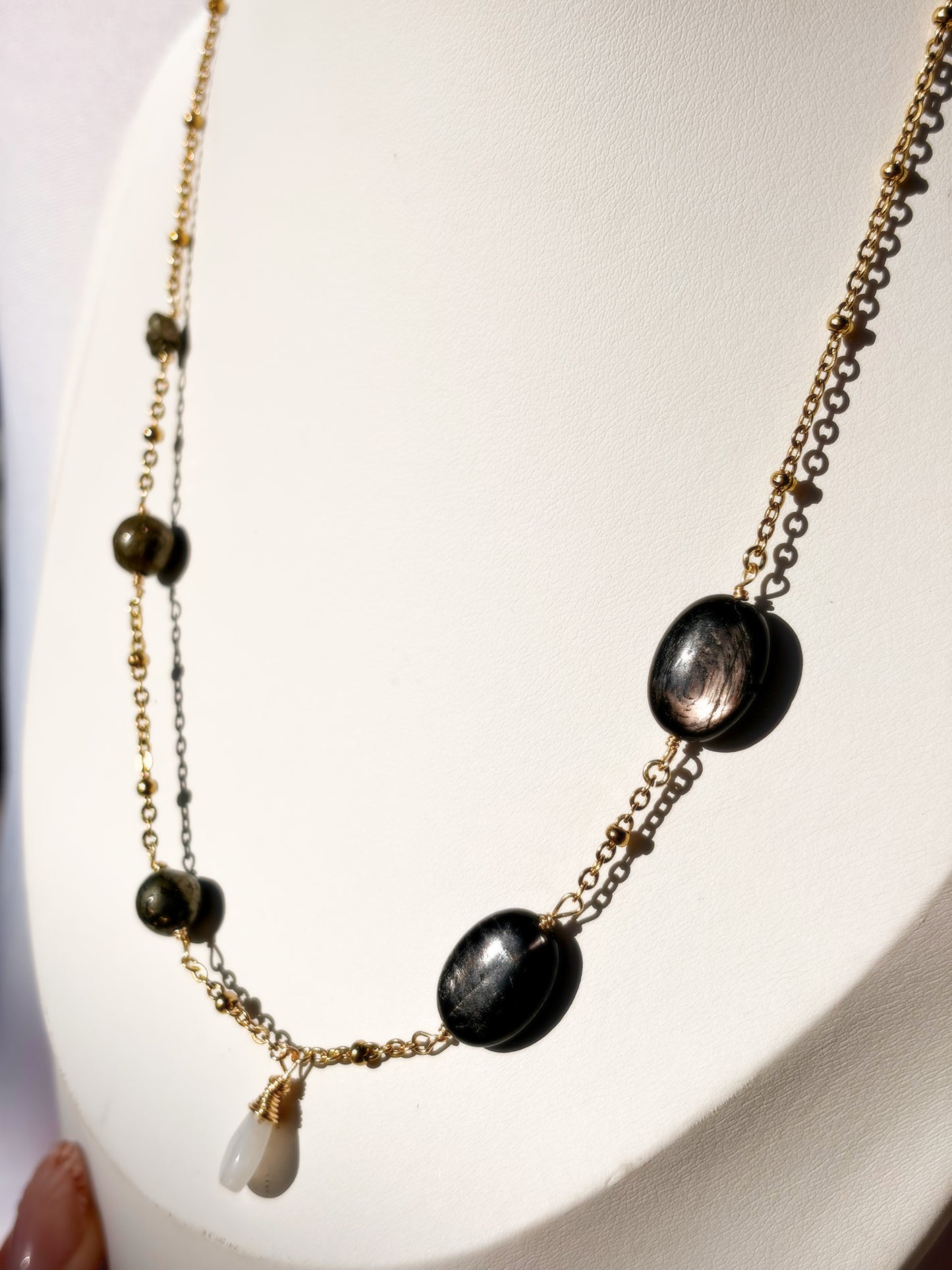 A gold-toned layered necklace displayed on a white jewelry bust. The necklace features a delicate gold chain with dark metallic oval HYPERSTHENE, Pyrite round beads, and a small white Rainbow Moonstone teardrop charm.