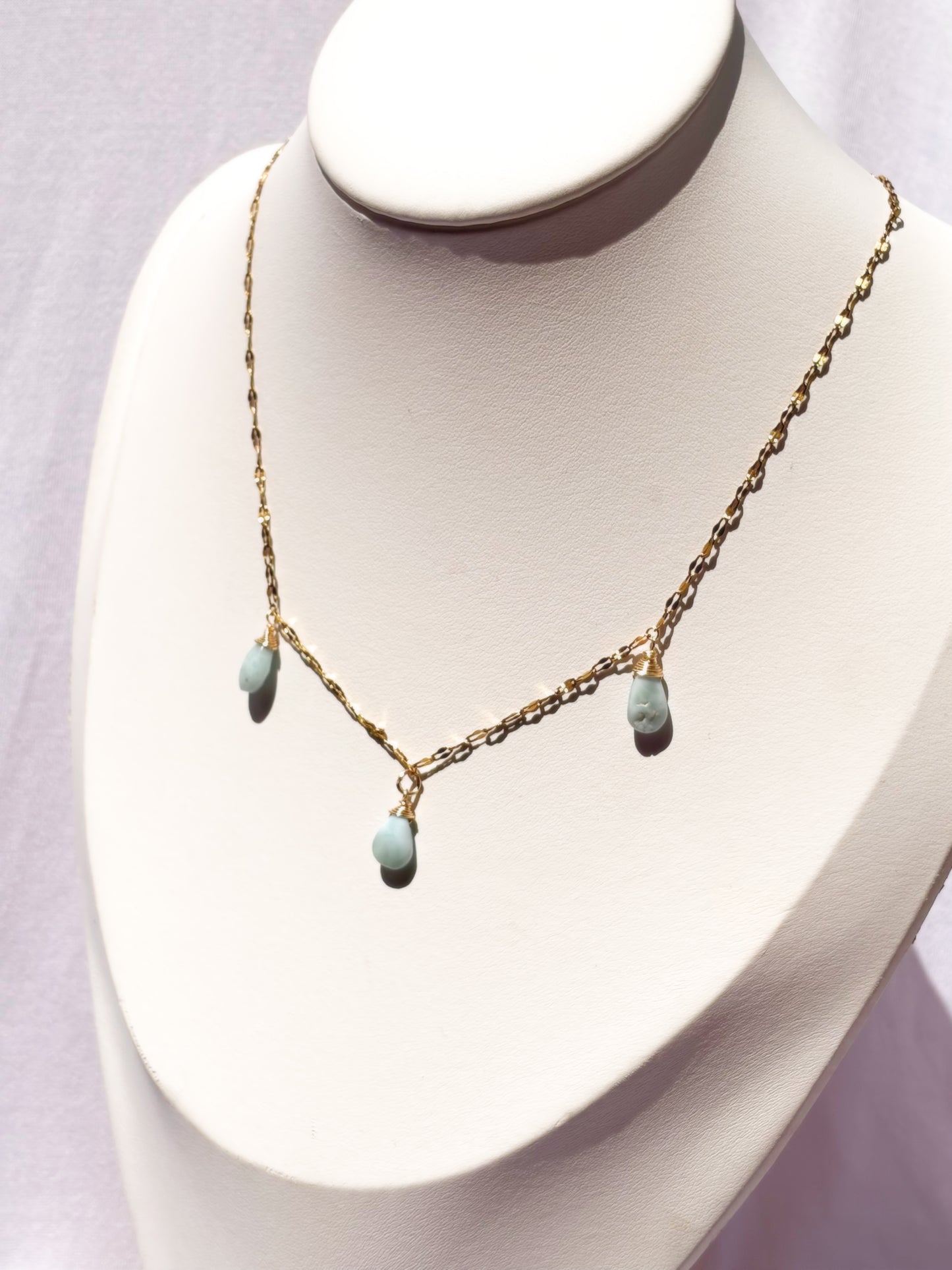 A delicate gold chain necklace with three small light blue teardrop Larimar beads, displayed on a white jewelry bust.