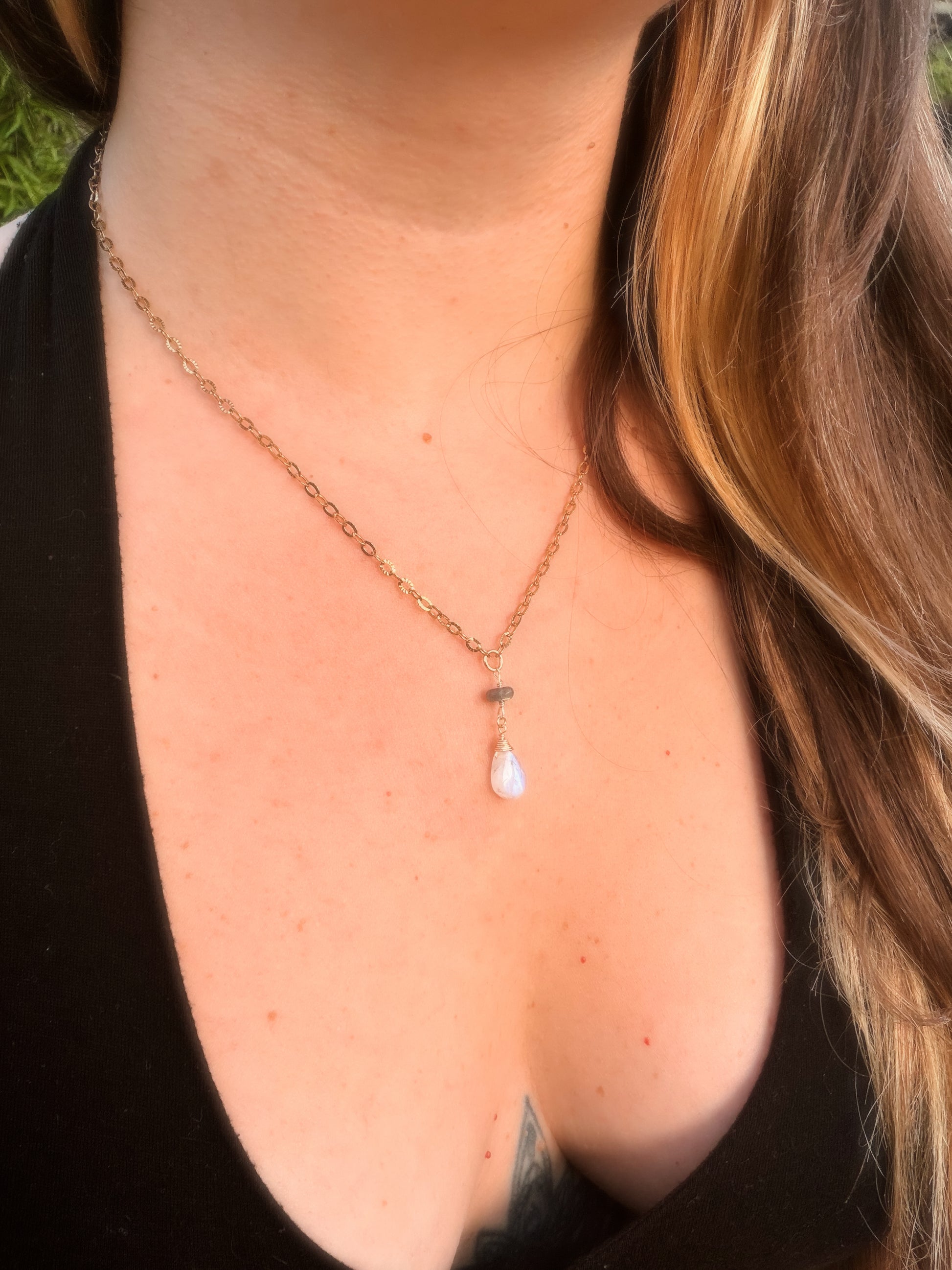 Close-up of a rainbow moonstone and labradorite stainless steel gold necklace with a teardrop pendant on a person wearing a black top.